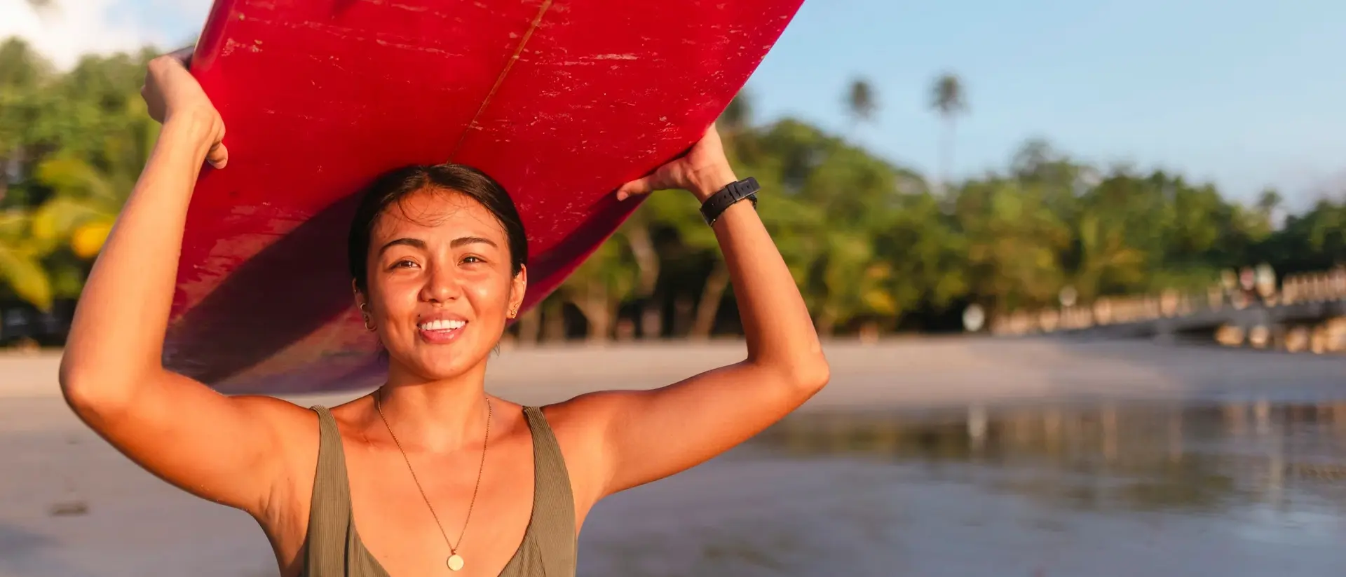 A smiling woman holds a red surfboard overhead at the beach, with palm trees and water in the background.