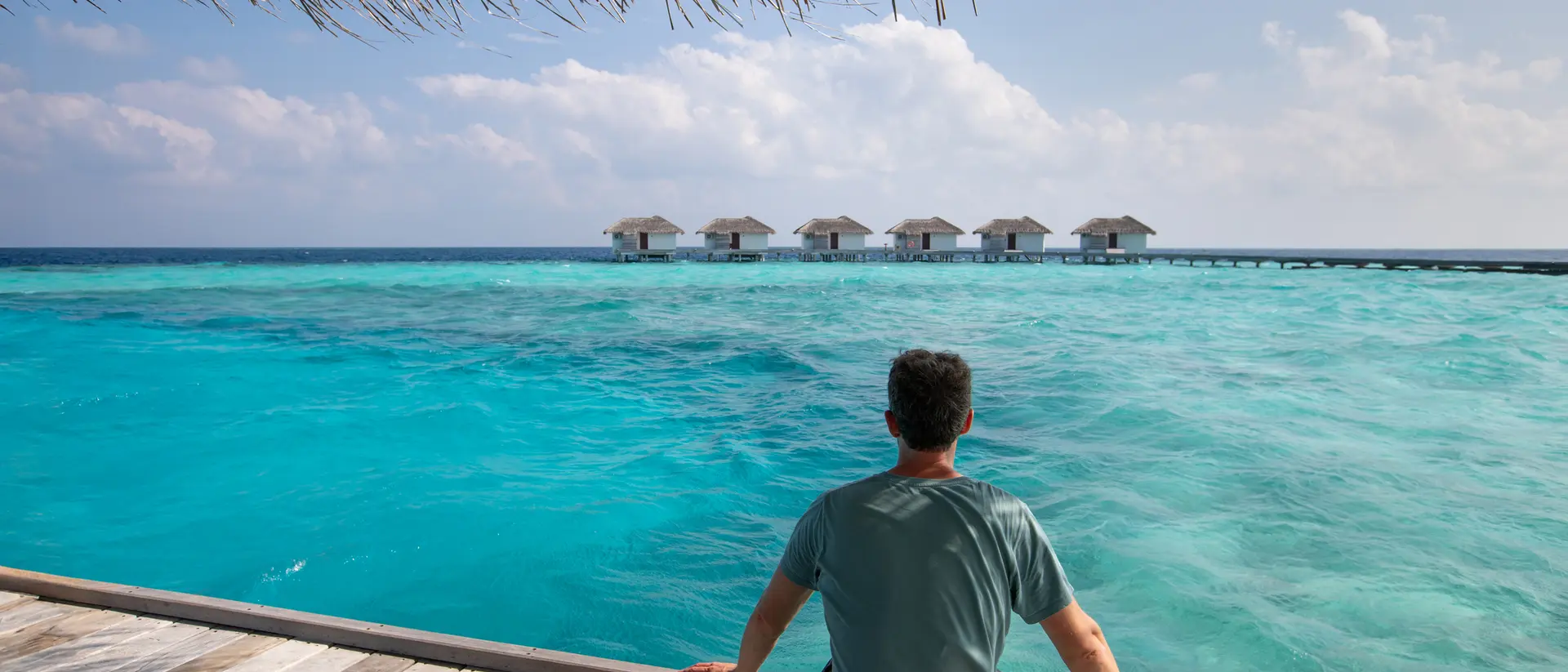 Person sitting on a wooden deck overlooking a vast turquoise sea and a row of overwater bungalows under a blue sky.