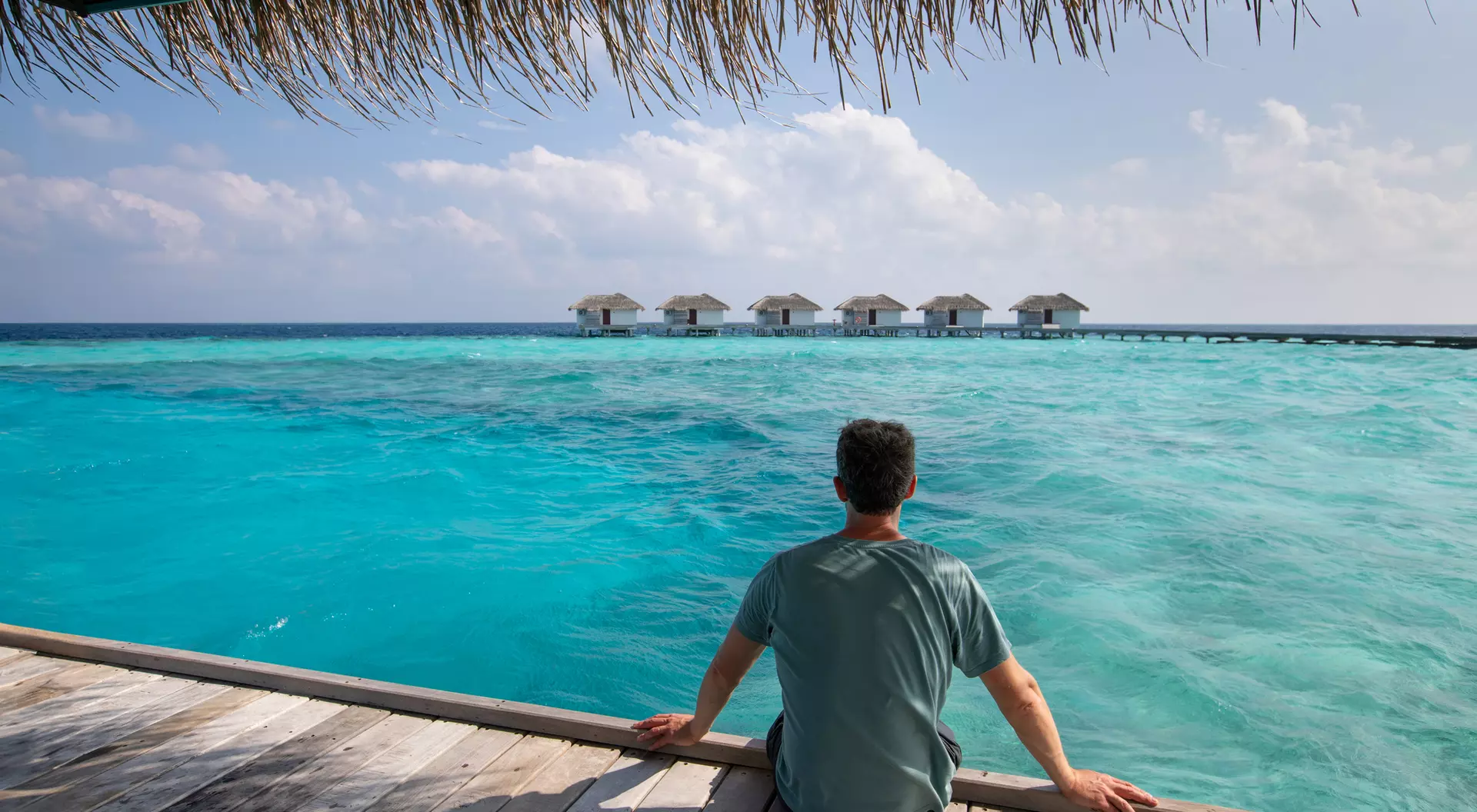 Person sitting on a wooden deck overlooking a vast turquoise sea and a row of overwater bungalows under a blue sky.