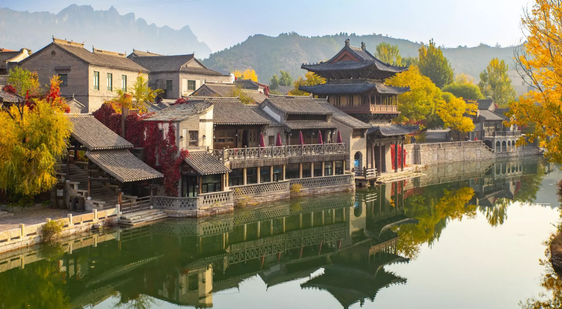 Scenic waterfront view of traditional buildings with autumn foliage mirrored in calm water. Mountains in the background.