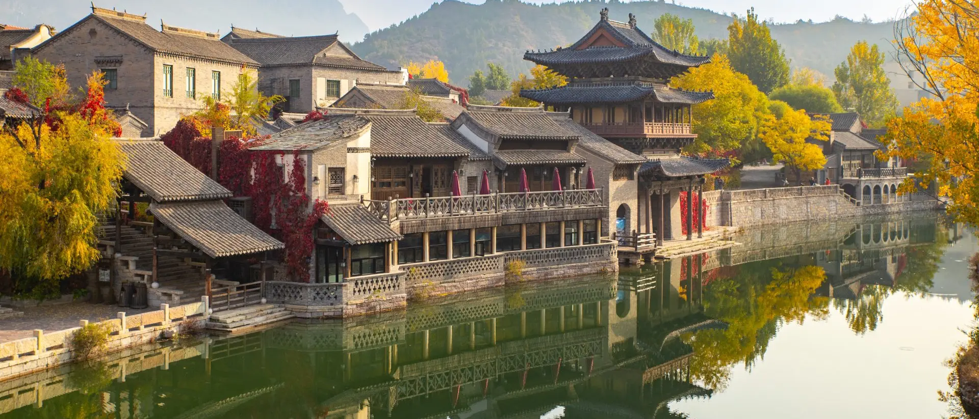Scenic waterfront view of traditional buildings with autumn foliage mirrored in calm water. Mountains in the background.