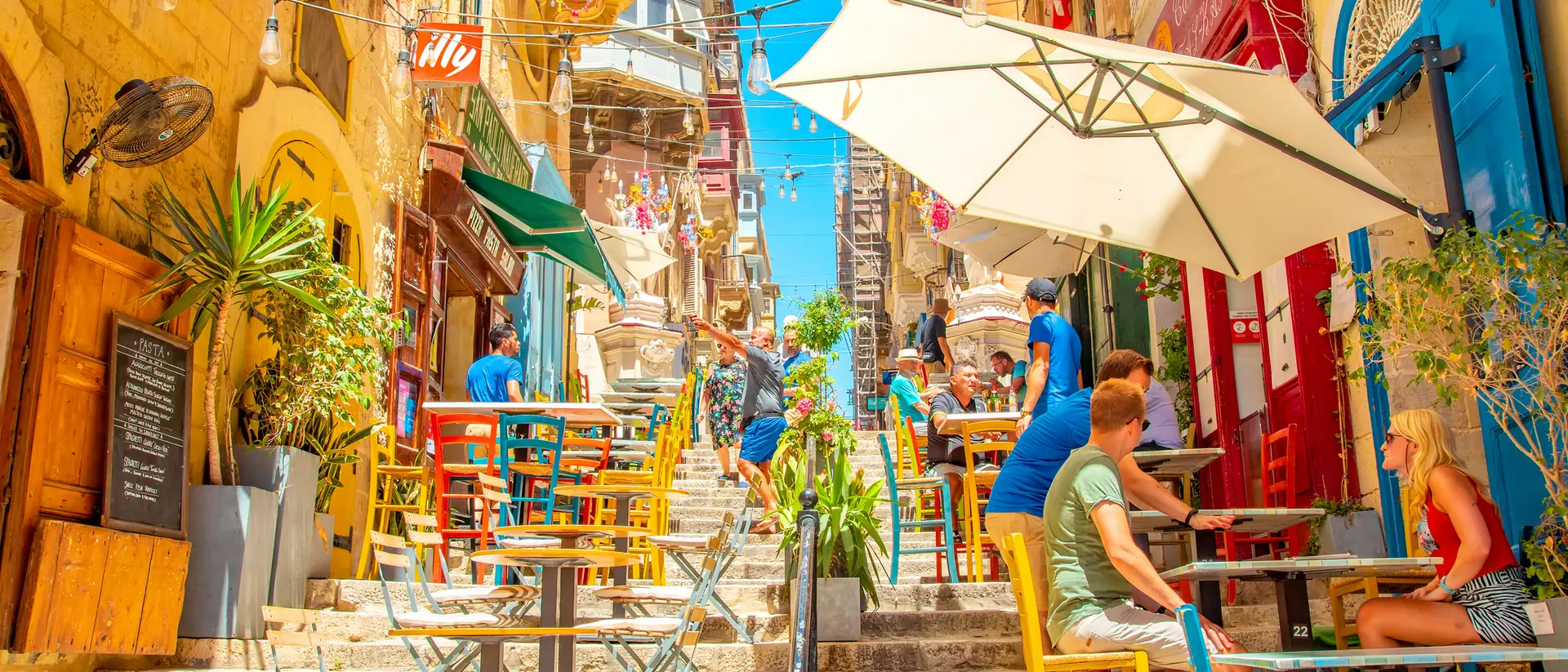 Colorful outdoor cafes line a sunlit, vibrant street, with people enjoying drinks and a bright blue sky overhead.