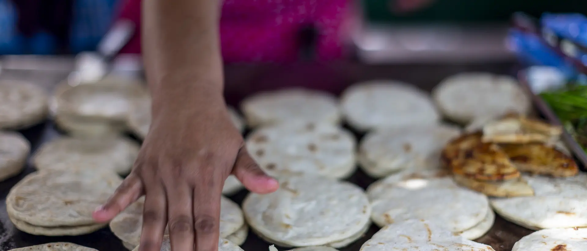 A hand spreads butter on freshly made flatbreads on a cooking surface, surrounded by more flatbreads and some grilled food.