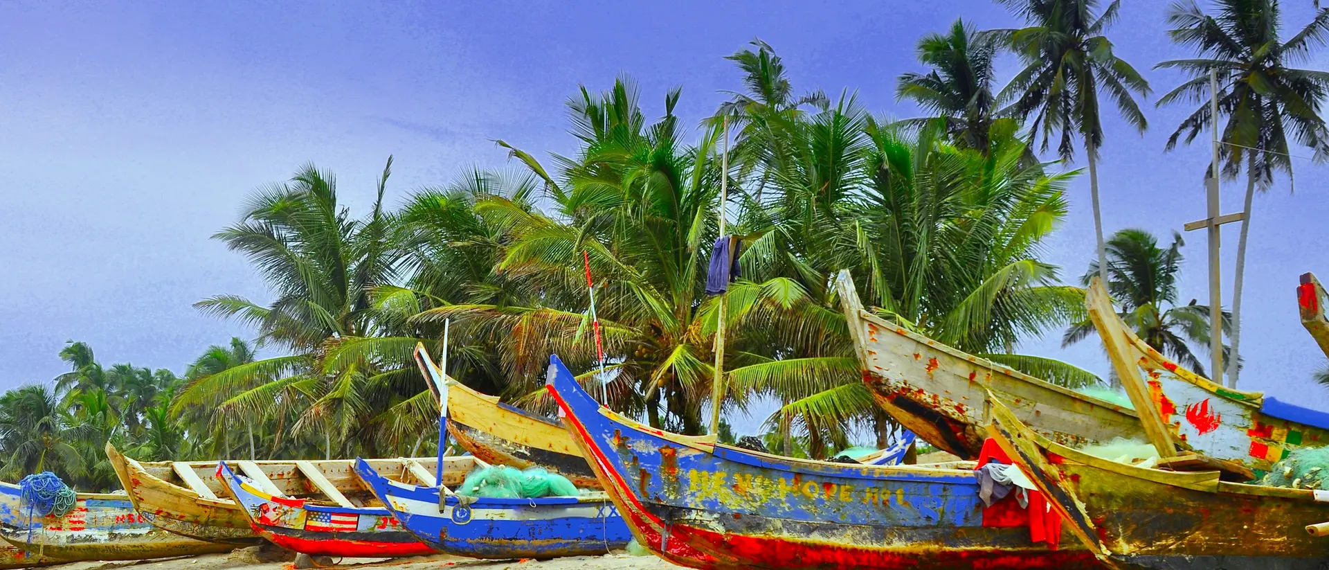 Colorful fishing boats lined up on a sandy beach, surrounded by lush palm trees under a blue sky.
