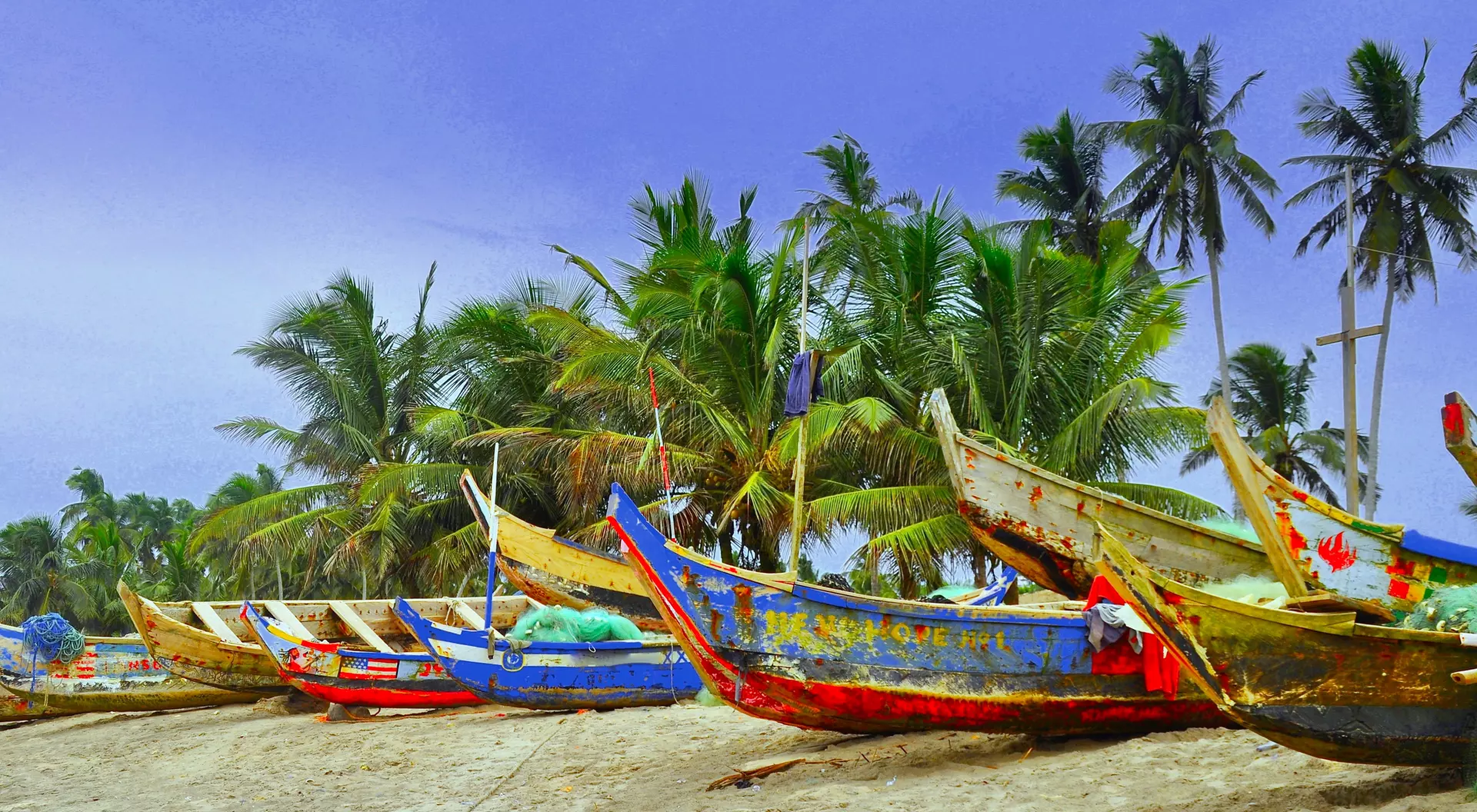 Colorful fishing boats lined up on a sandy beach, surrounded by lush palm trees under a blue sky.