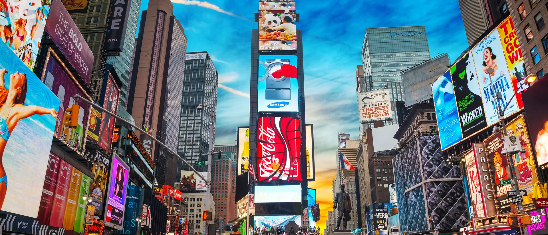 Crowds gather in Times Square, surrounded by vibrant billboards and a colorful sky at sunset.