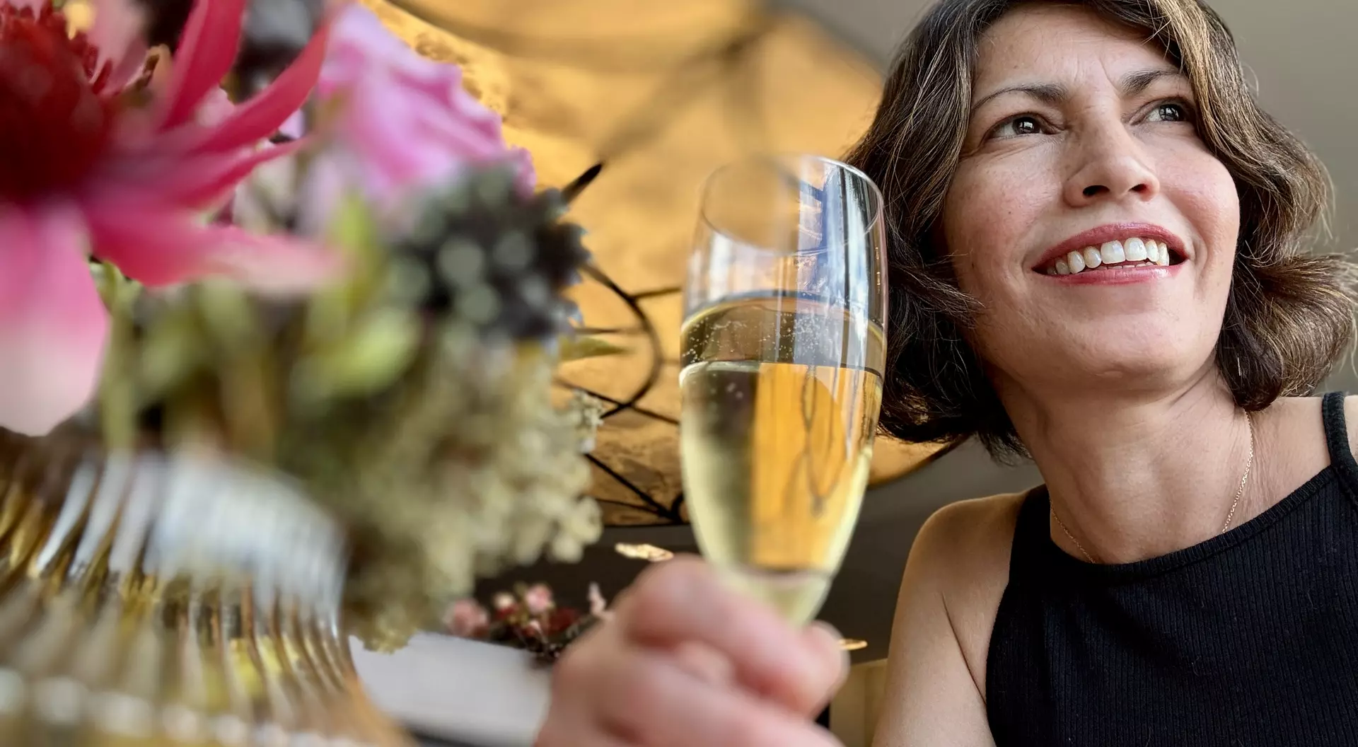 A smiling woman holding a glass of champagne, surrounded by colorful flowers in a bright, elegant setting.