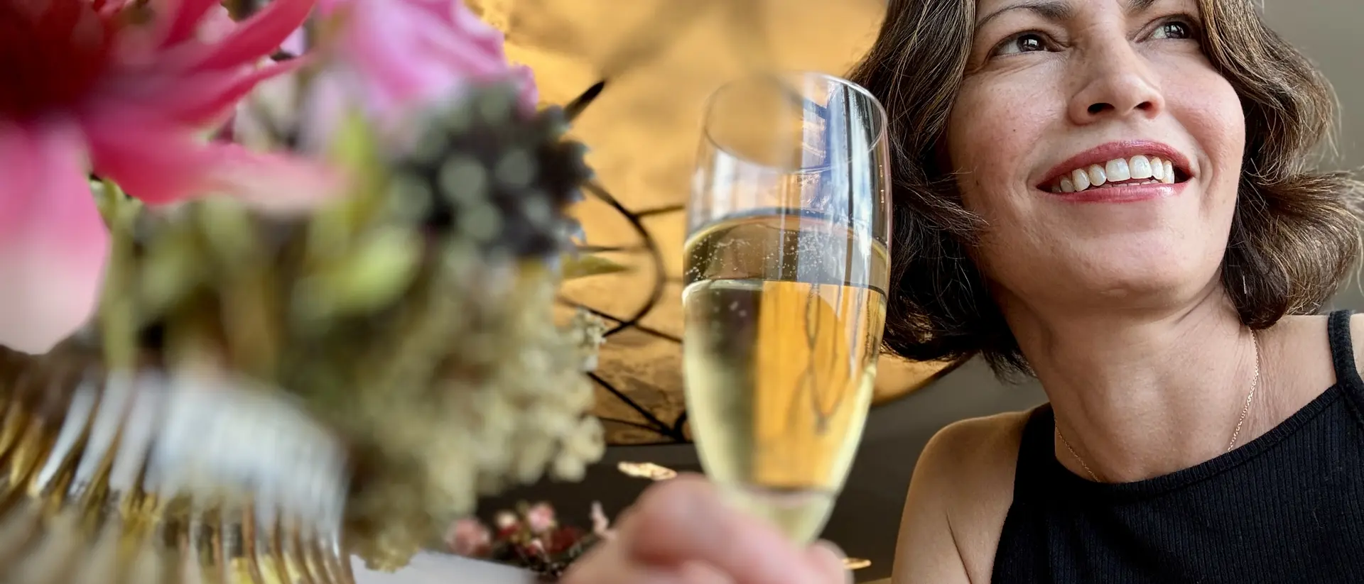 A smiling woman holding a glass of champagne, surrounded by colorful flowers in a bright, elegant setting.