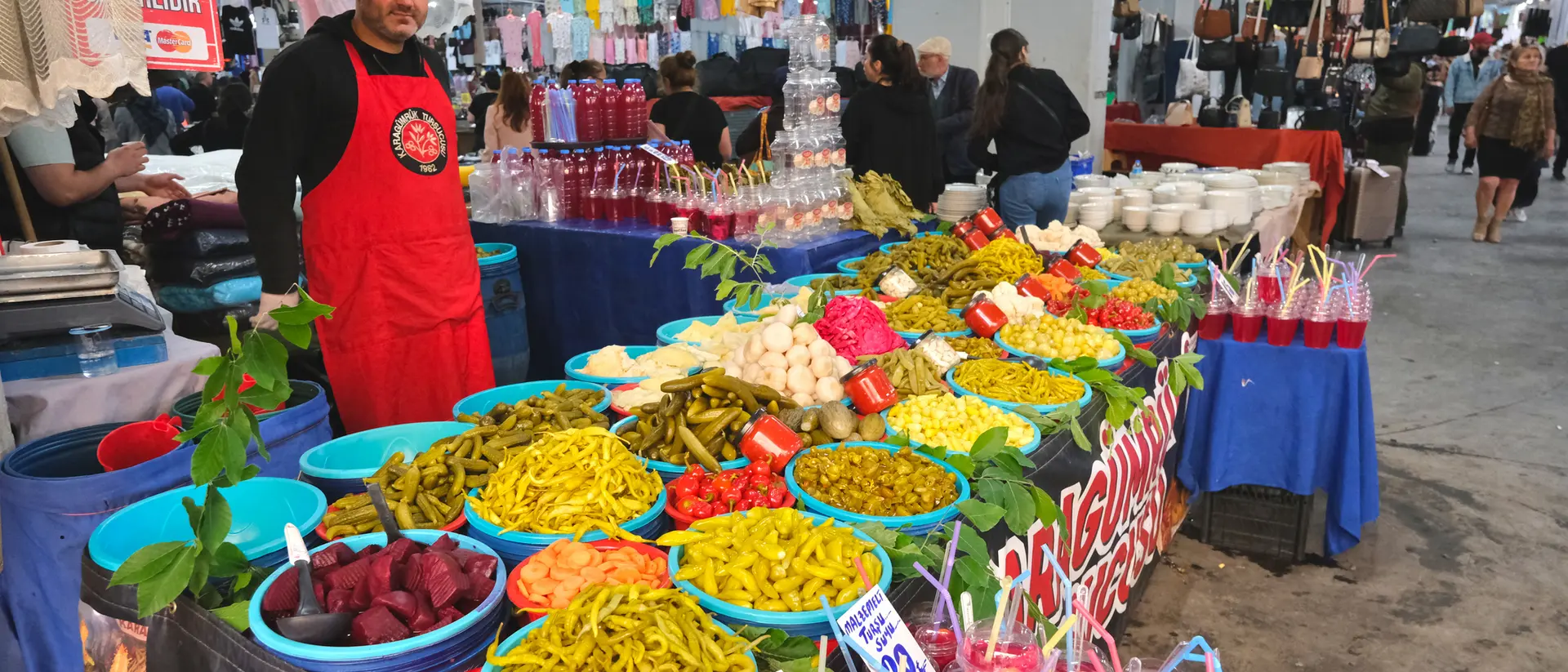 A vendor in an apron stands behind a vibrant table filled with colorful fruits, vegetables, and drinks at a market.