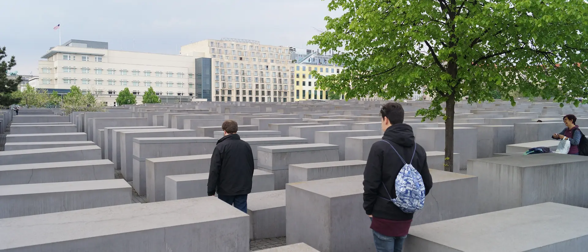 Visitors walk among tall concrete slabs at the Holocaust Memorial in Berlin, with a tree and buildings in the background.