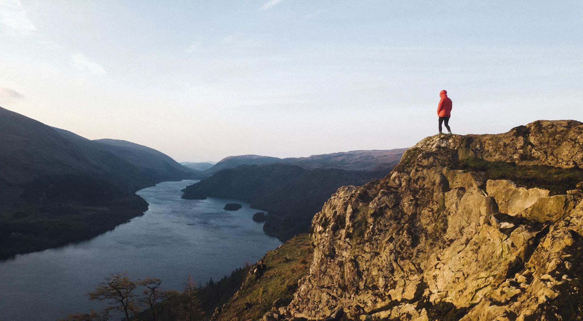 A person in a red jacket stands on a rocky outcrop overlooking a river and mountains at sunset.