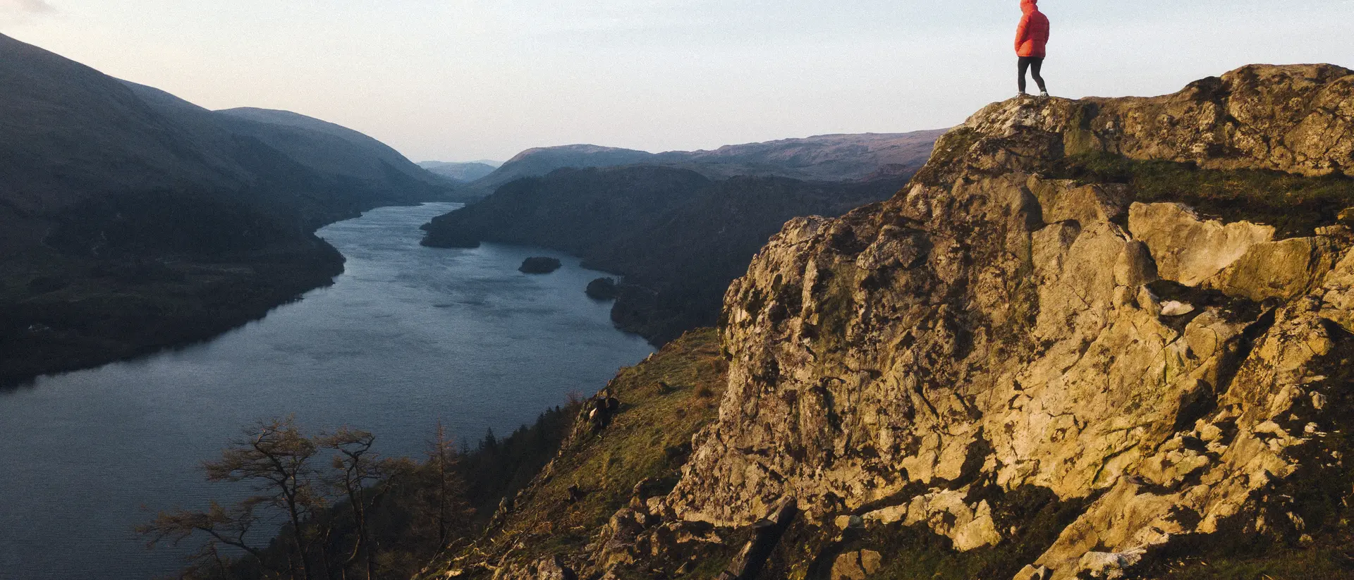 A person in a red jacket stands on a rocky outcrop overlooking a river and mountains at sunset.
