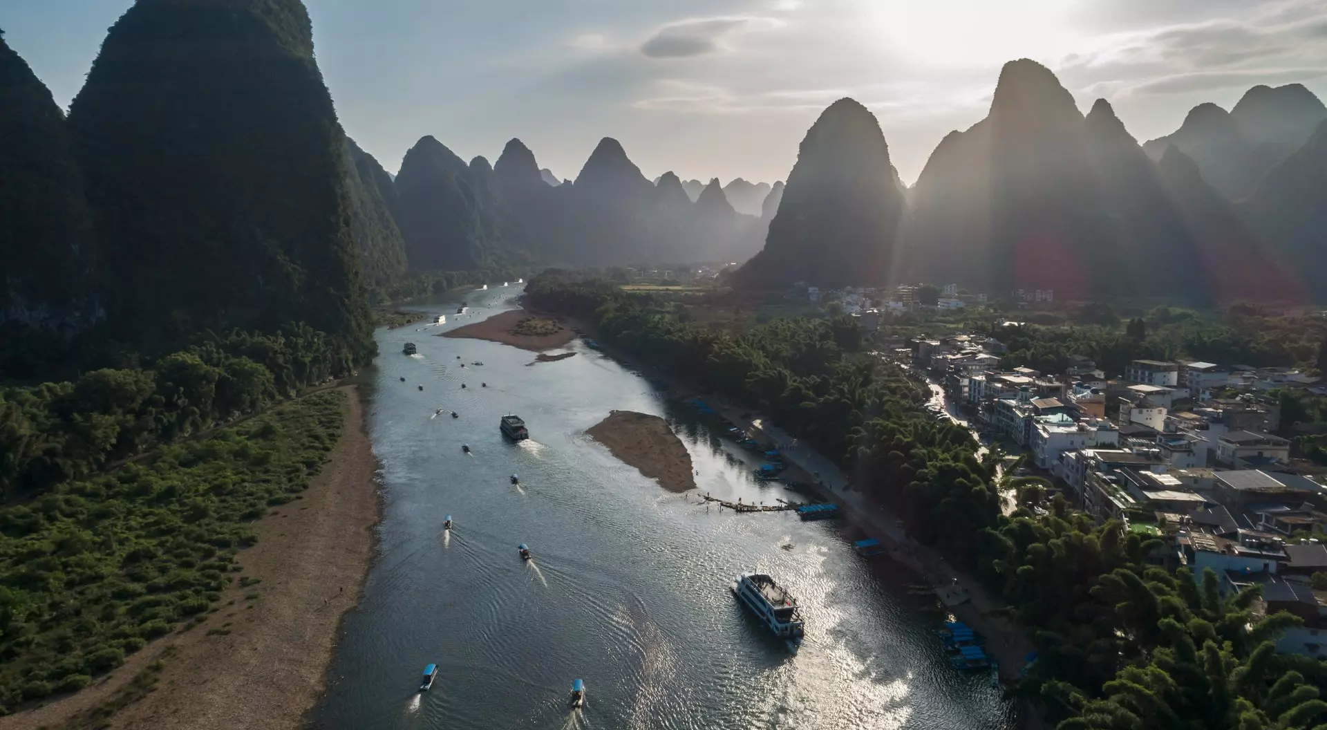 Aerial view of a river surrounded by lush mountains and a small town, with boats navigating the water in the foreground.