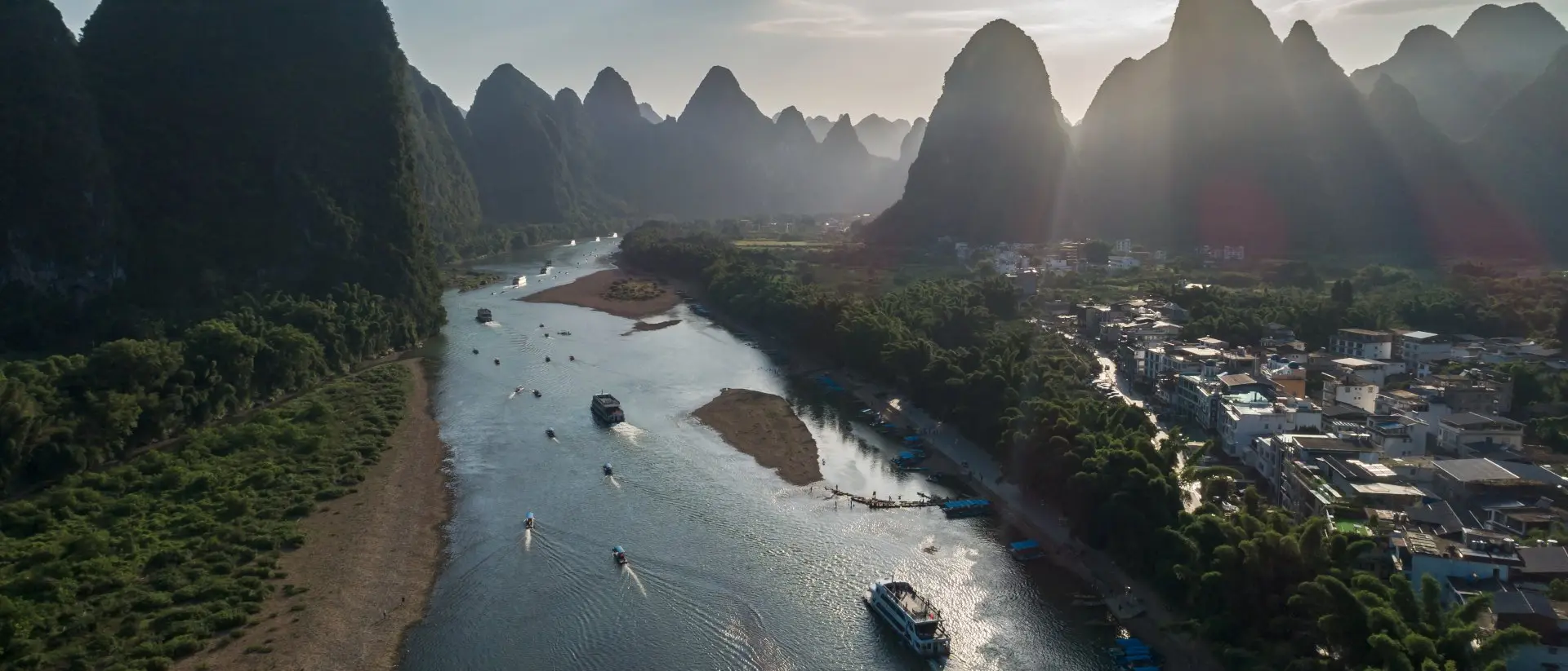 Aerial view of a river surrounded by lush mountains and a small town, with boats navigating the water in the foreground.