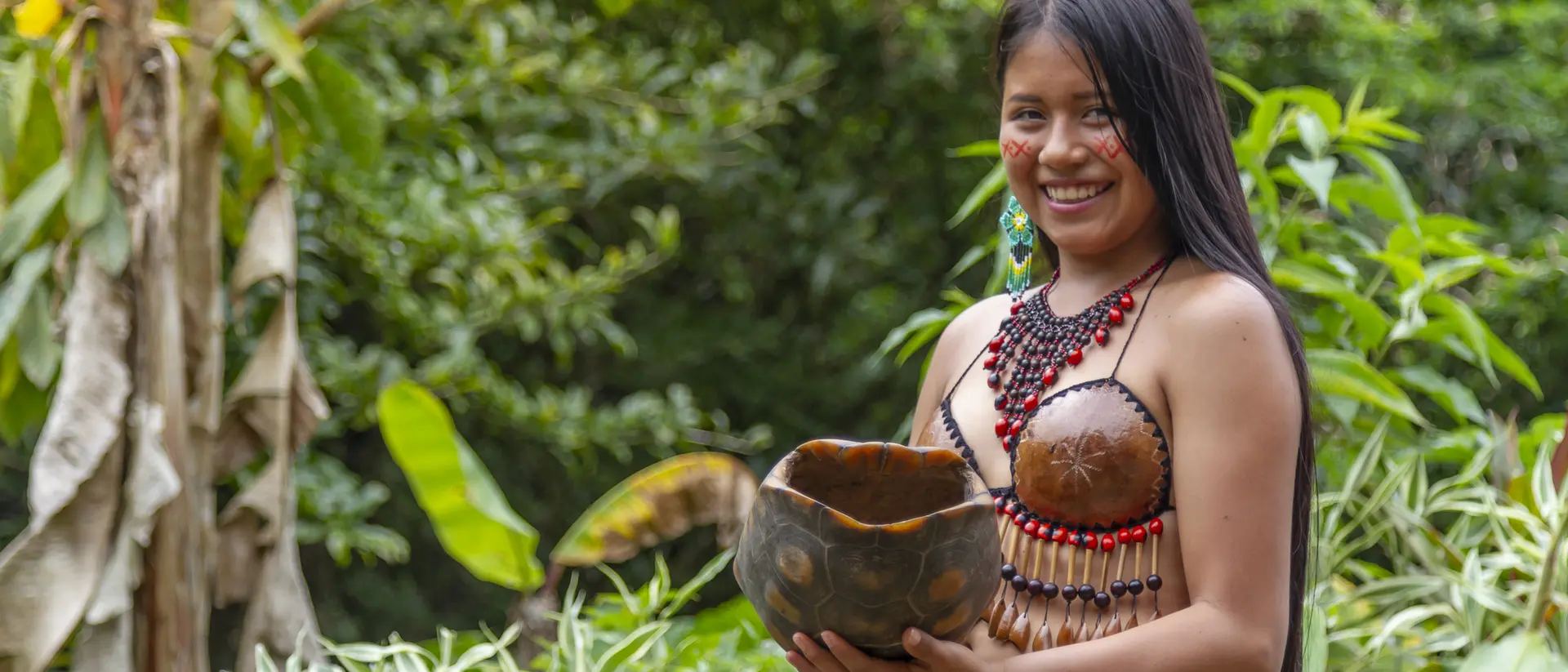 A woman in traditional attire smiles while holding a decorative bowl surrounded by lush greenery.