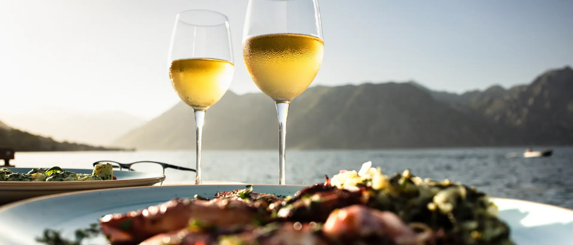 Two glasses of white wine in front of a scenic view with mountains, alongside a plate of seafood and greens.