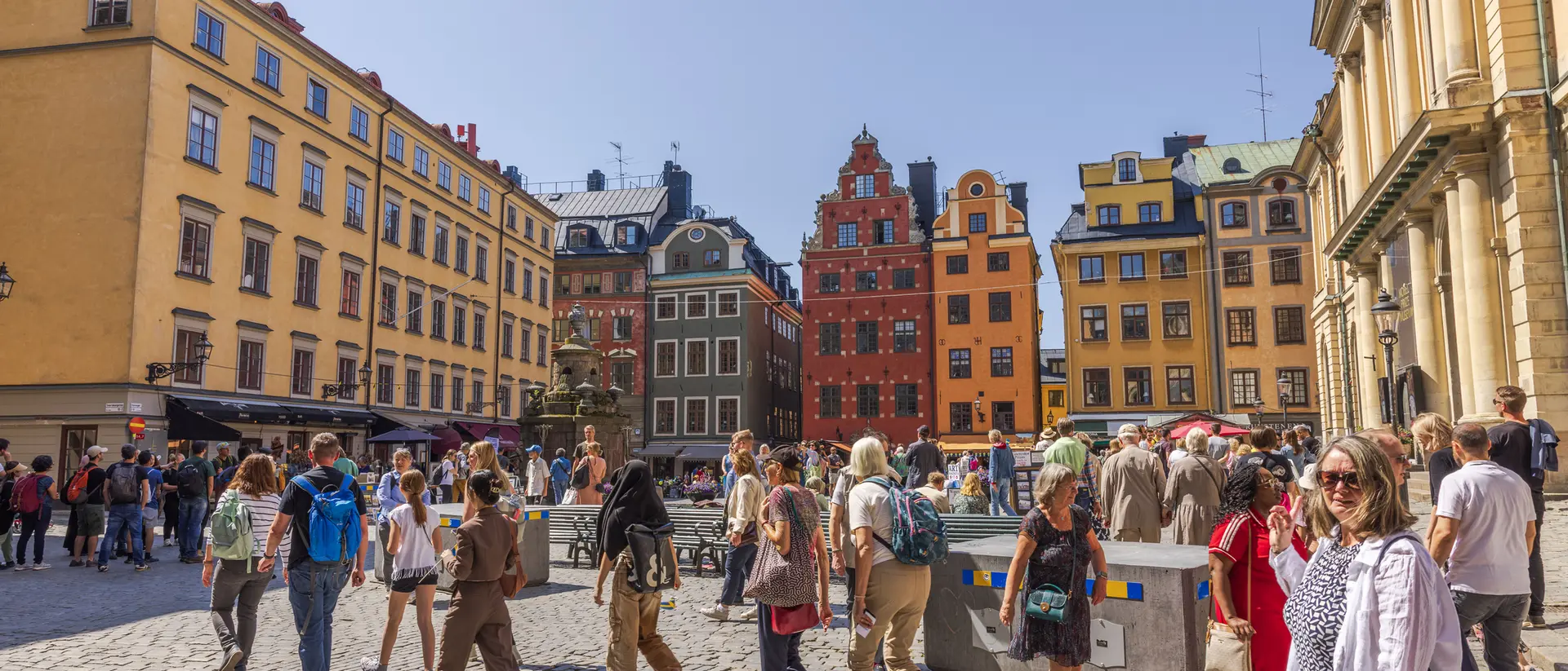 Crowded cobblestone square with people walking, surrounded by colorful historic buildings under a clear blue sky.