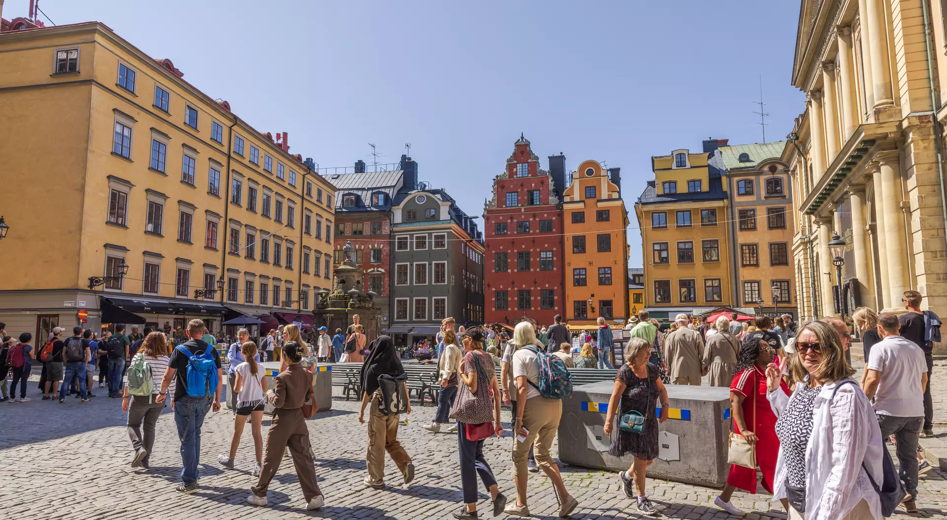 Crowded cobblestone square with people walking, surrounded by colorful historic buildings under a clear blue sky.