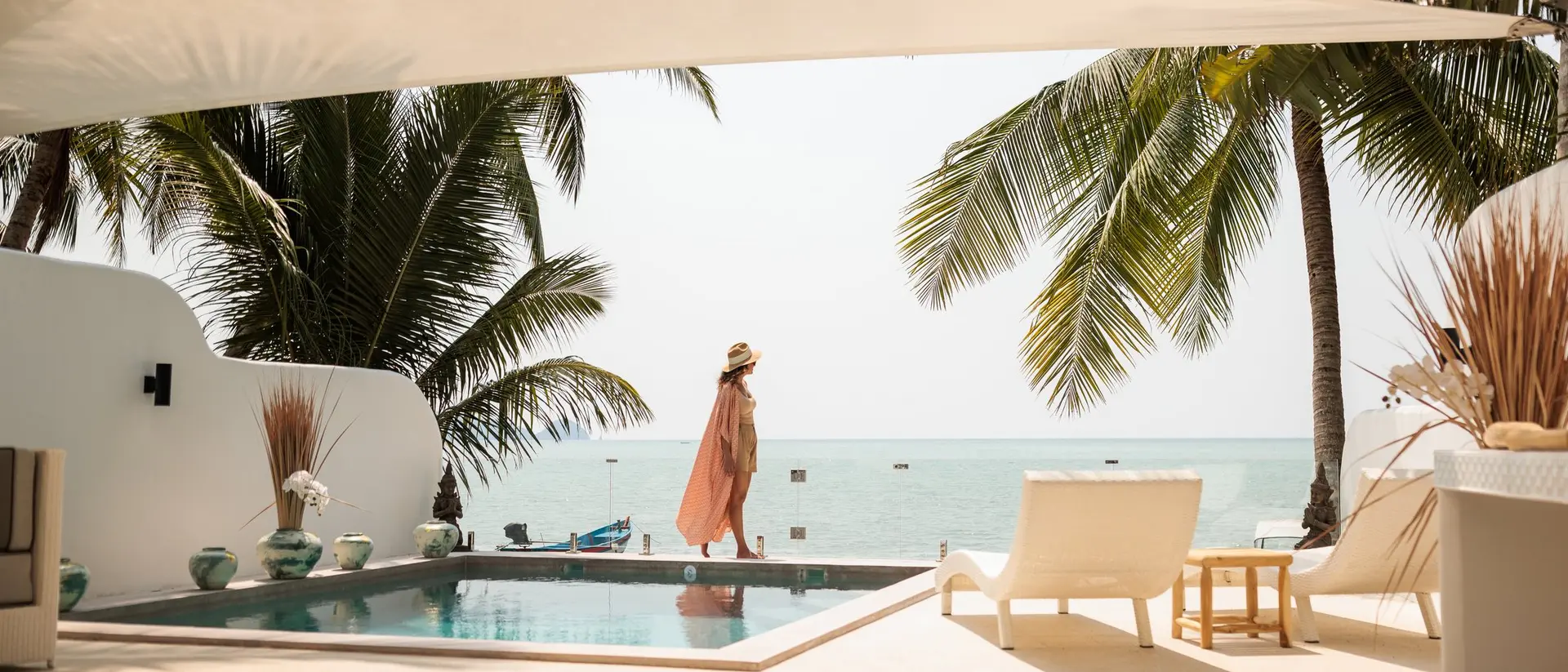 A woman in a pink cover-up stands by a pool, overlooking the ocean surrounded by palm trees and a sunny sky.