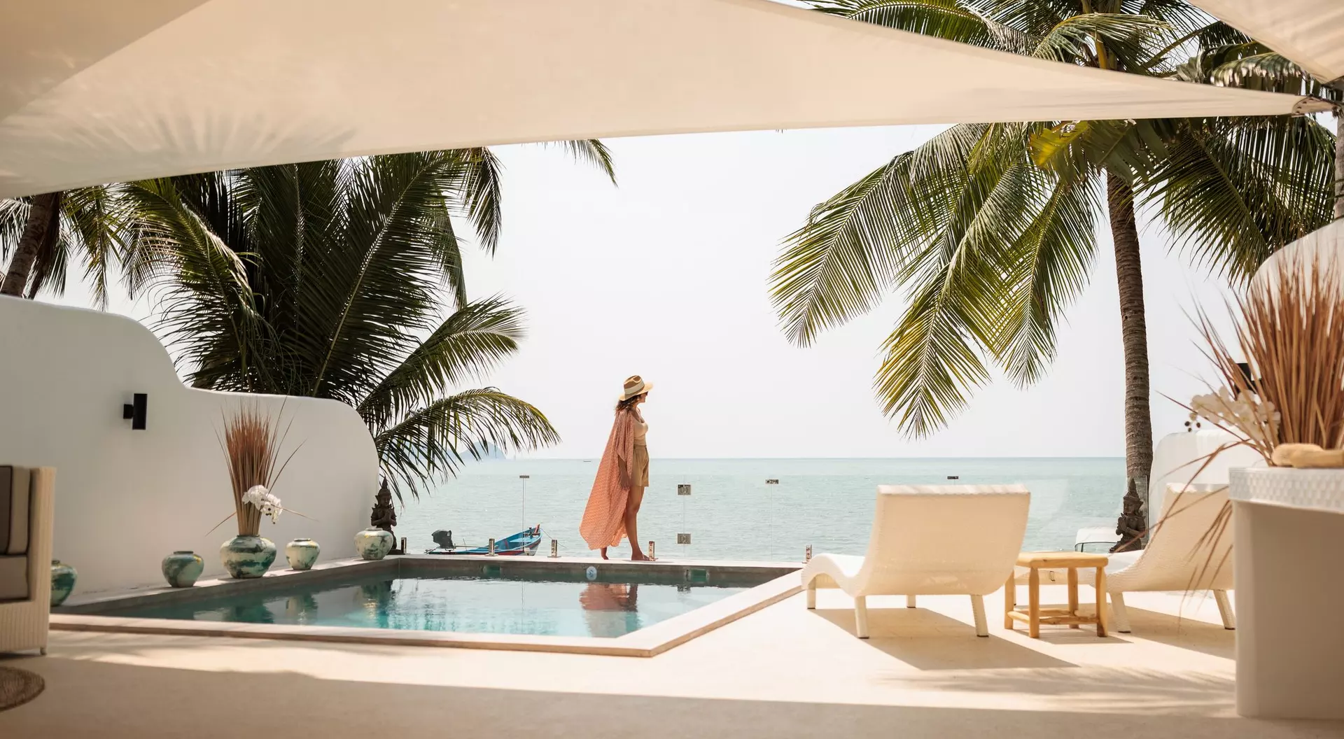 A woman in a pink cover-up stands by a pool, overlooking the ocean surrounded by palm trees and a sunny sky.