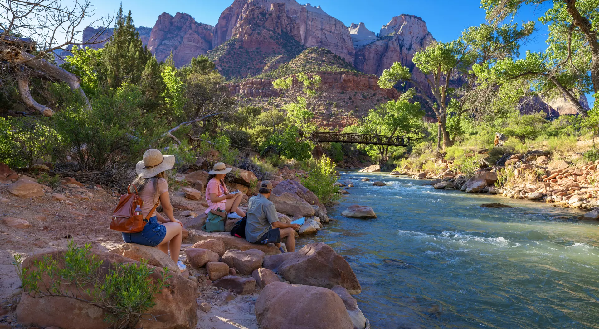 People sit by a river in a canyon, surrounded by trees and mountains on a sunny day.
