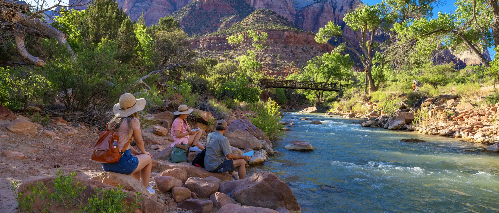 People sit by a river in a canyon, surrounded by trees and mountains on a sunny day.