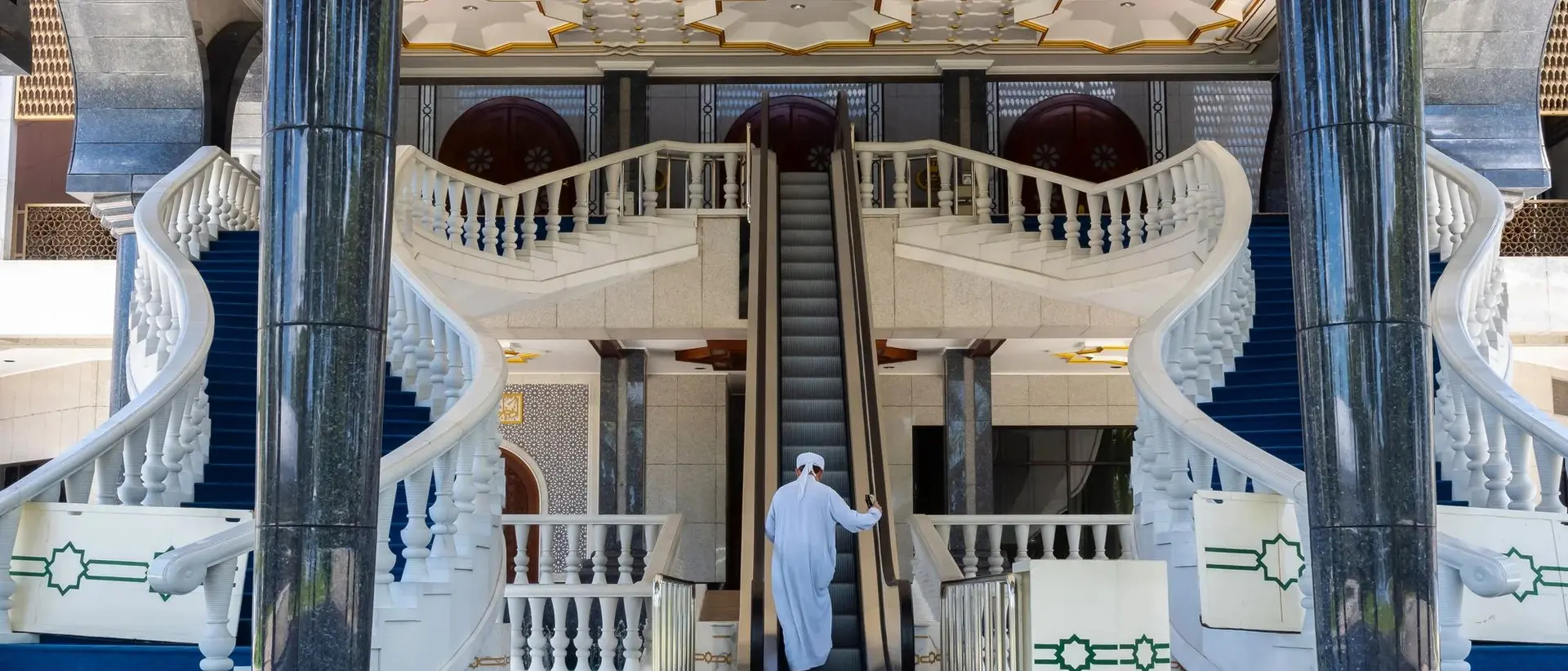 A person in traditional attire stands on an escalator in an ornate hall with grand staircases and a beautifully decorated ceiling.