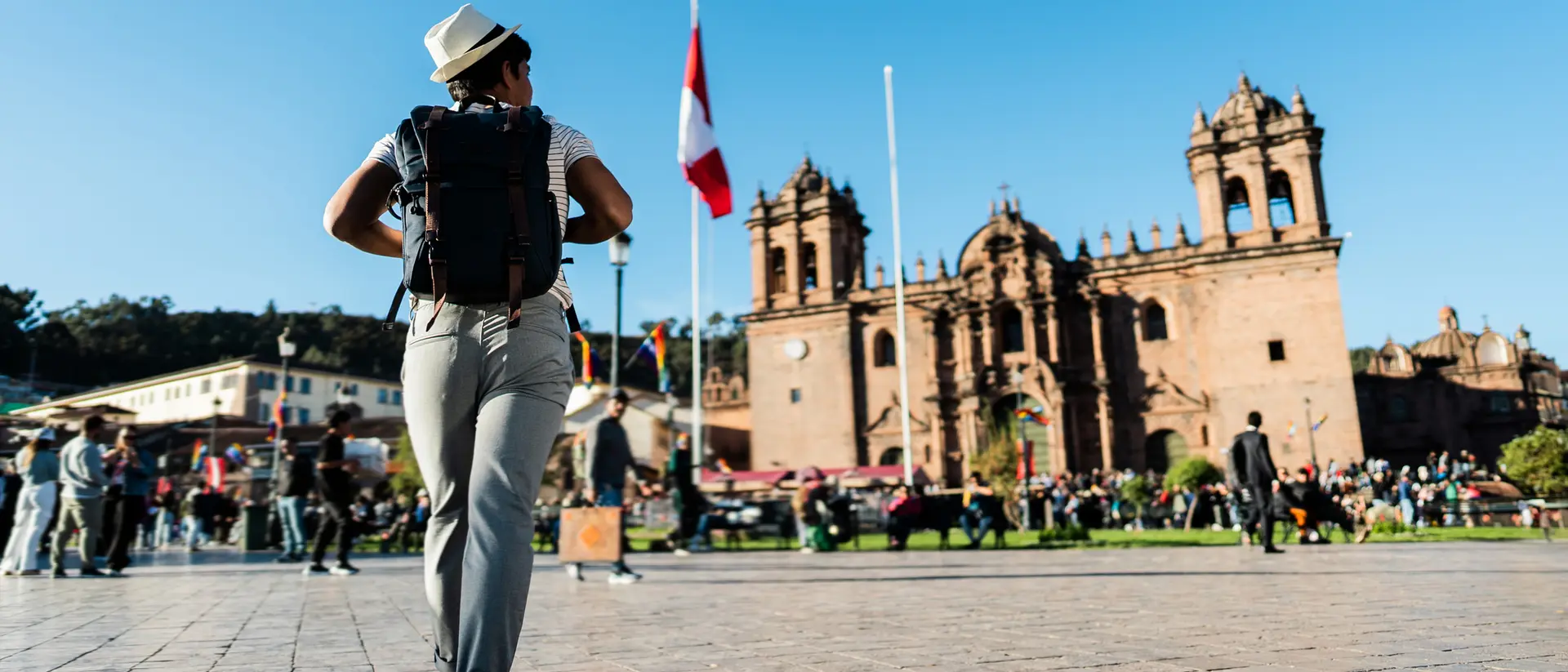 A person in a hat walks through a bustling plaza with historic buildings and flags in the background. Bright blue sky.