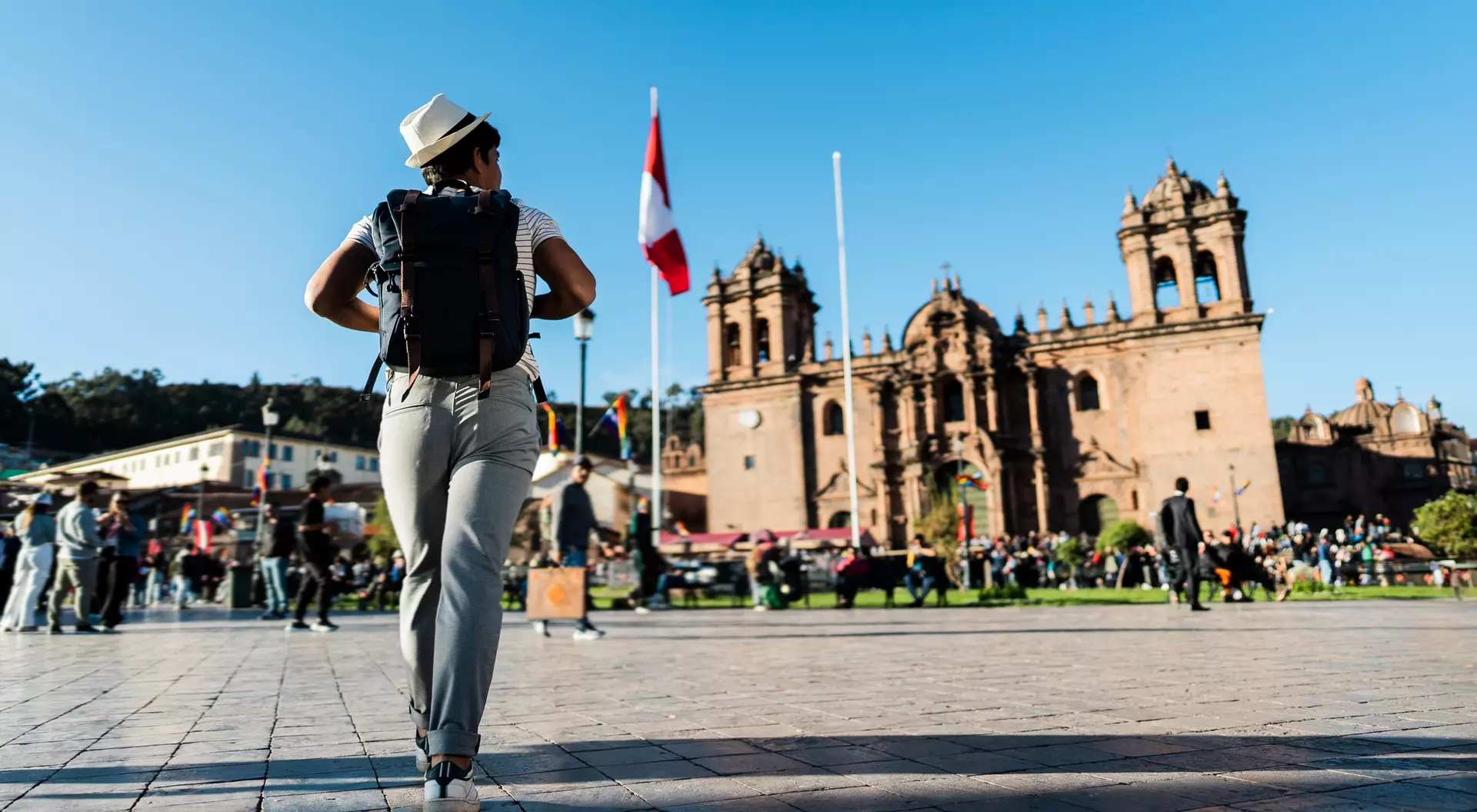 A person in a hat walks through a bustling plaza with historic buildings and flags in the background. Bright blue sky.