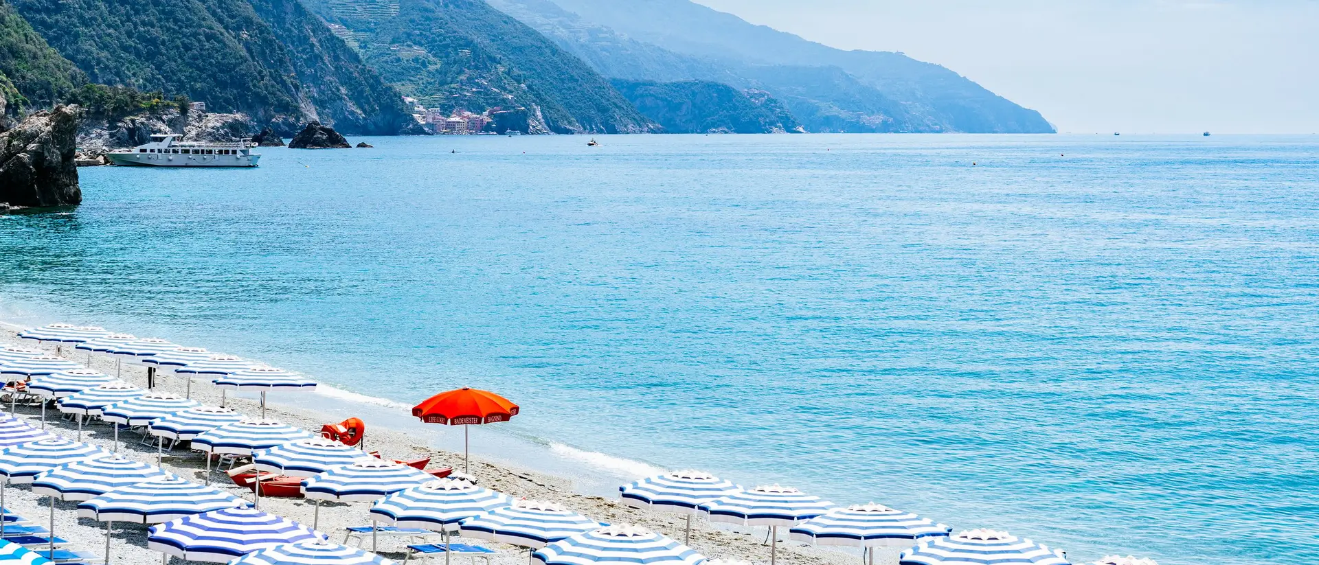 Beach with striped umbrellas and a red umbrella, overlooking calm turquoise waters and mountains in the background.