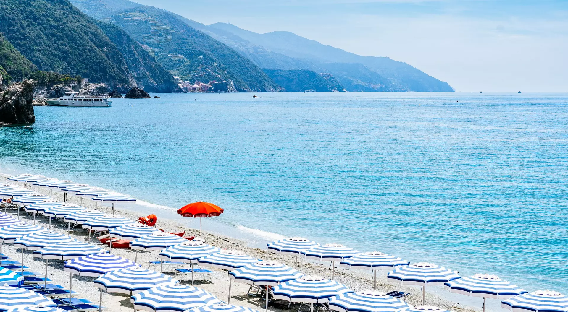 Beach with striped umbrellas and a red umbrella, overlooking calm turquoise waters and mountains in the background.