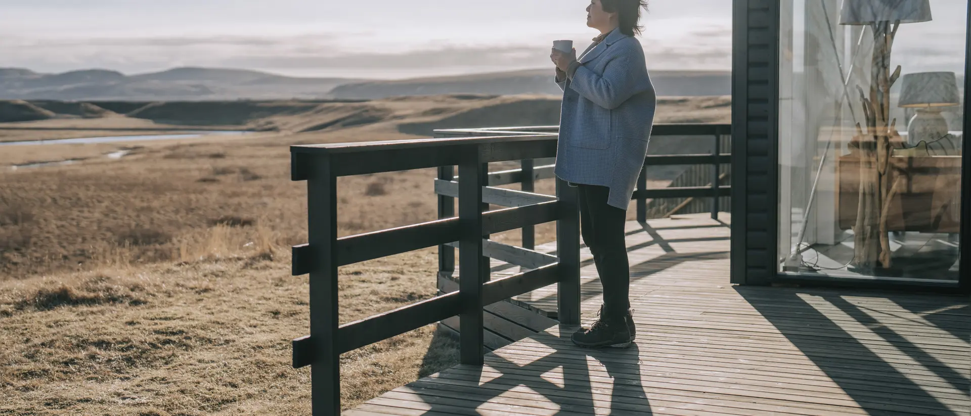 A person stands on a wooden deck, holding a cup, gazing at a scenic landscape of fields and distant hills.