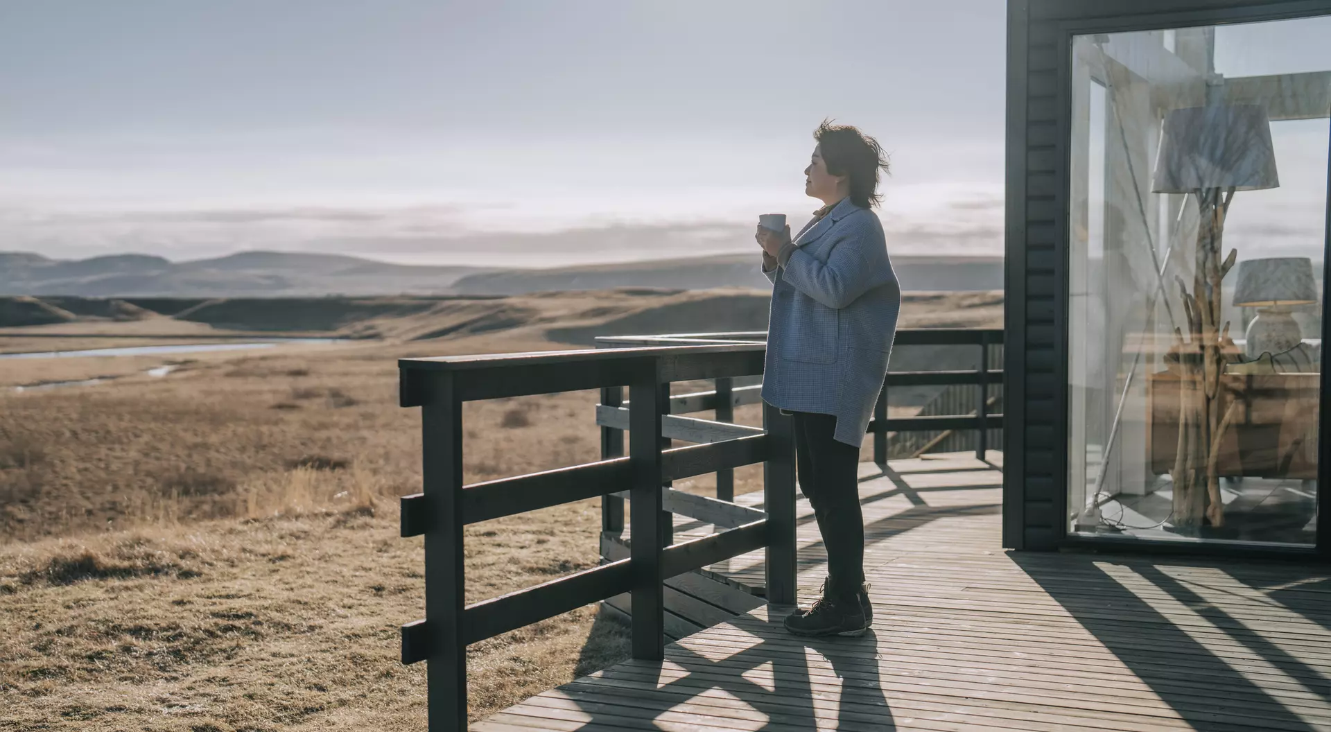 A person stands on a wooden deck, holding a cup, gazing at a scenic landscape of fields and distant hills.