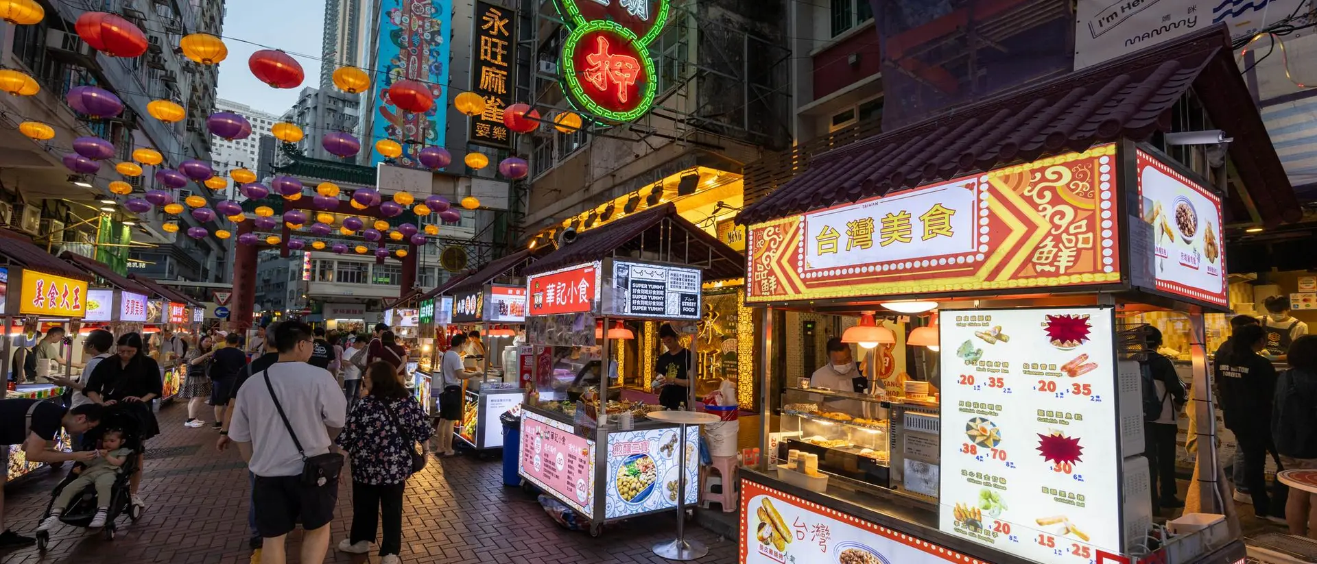 Vibrant street market scene with food stalls, colorful lanterns overhead, and people walking among the stalls.