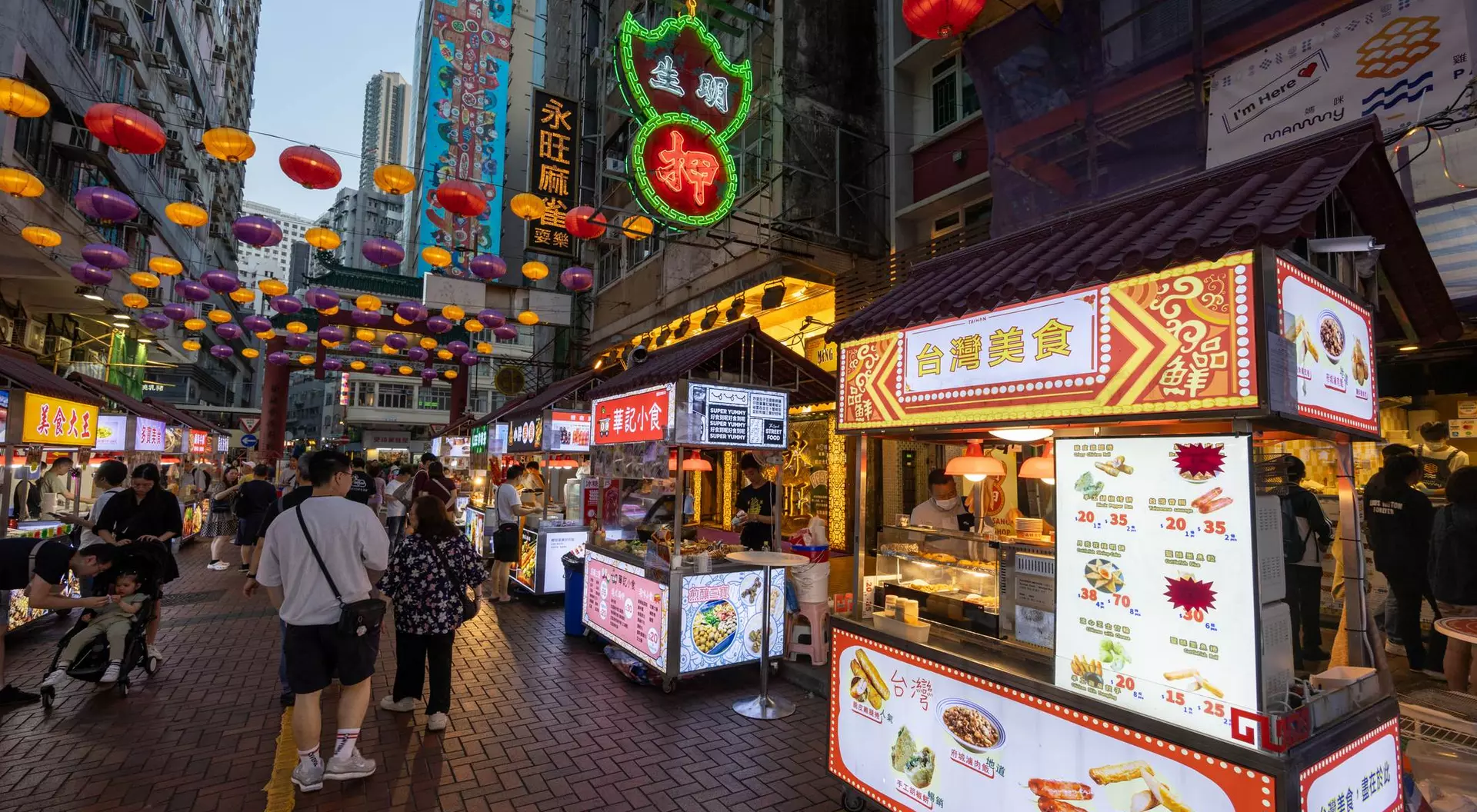 Vibrant street market scene with food stalls, colorful lanterns overhead, and people walking among the stalls.