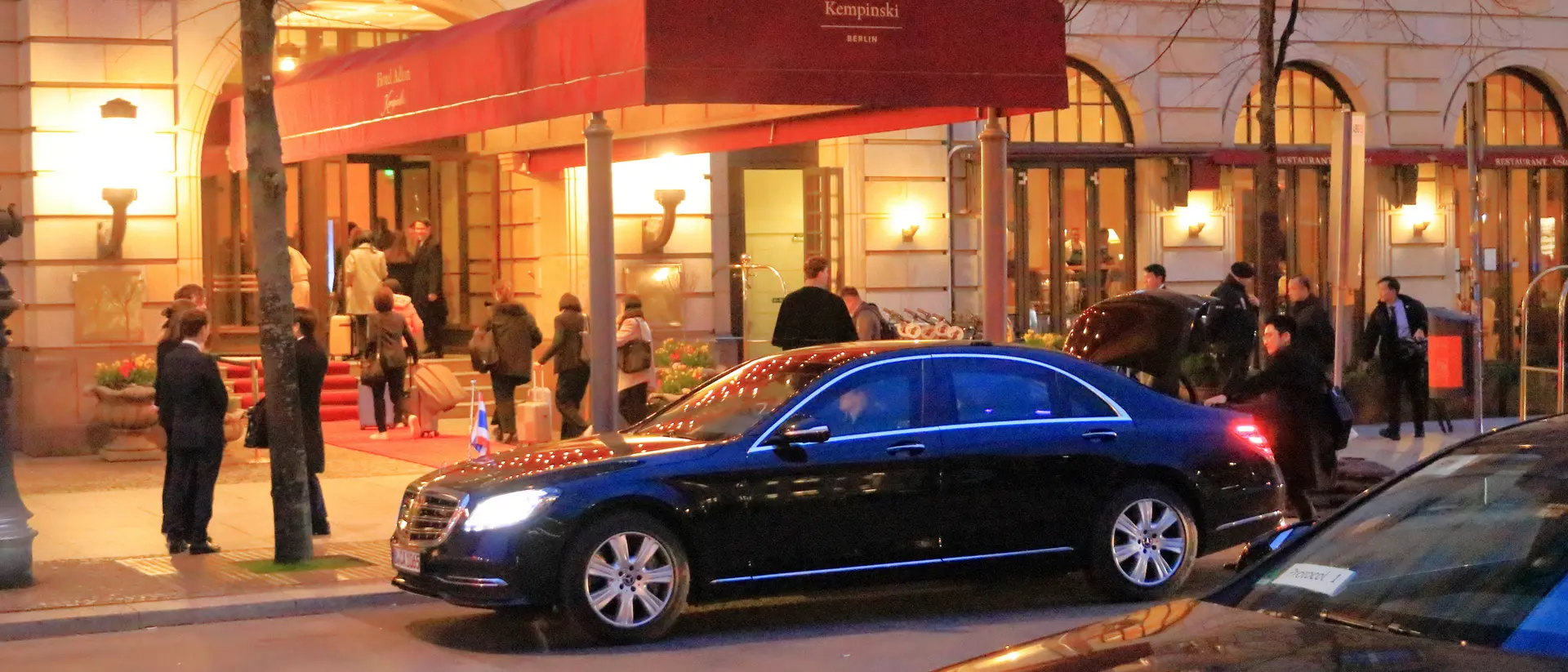 A black car parked in front of Hotel Adlon, with guests arriving under a red awning in the evening.