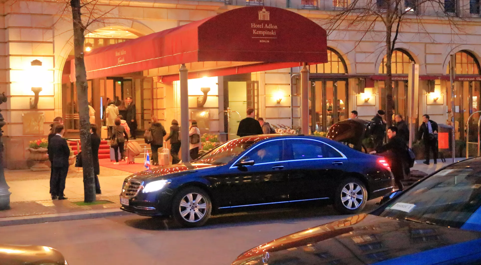 A black car parked in front of Hotel Adlon, with guests arriving under a red awning in the evening.