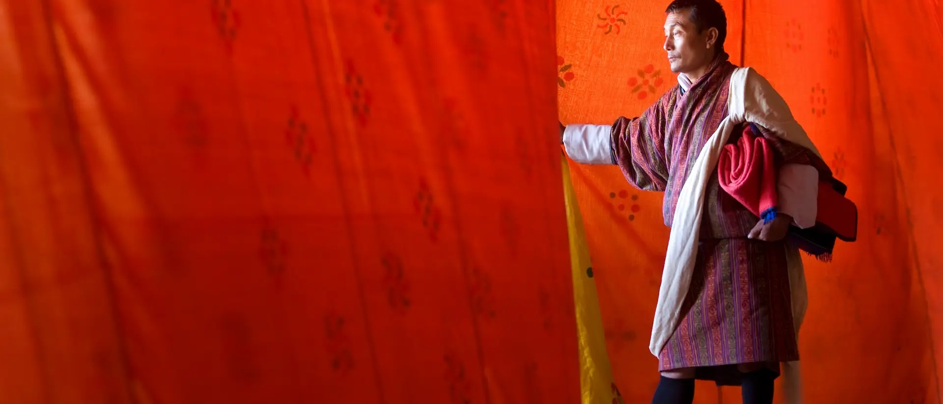 A man in traditional attire stands near colorful fabric, reaching out as he appears to enter a bright space.