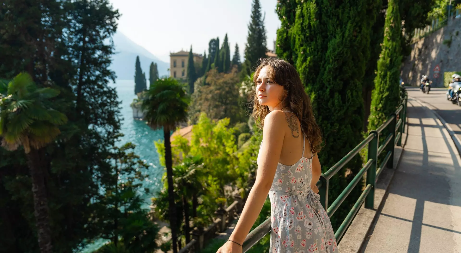 A woman in a floral dress leans on a railing, enjoying a sunny view of a lake surrounded by greenery and trees.