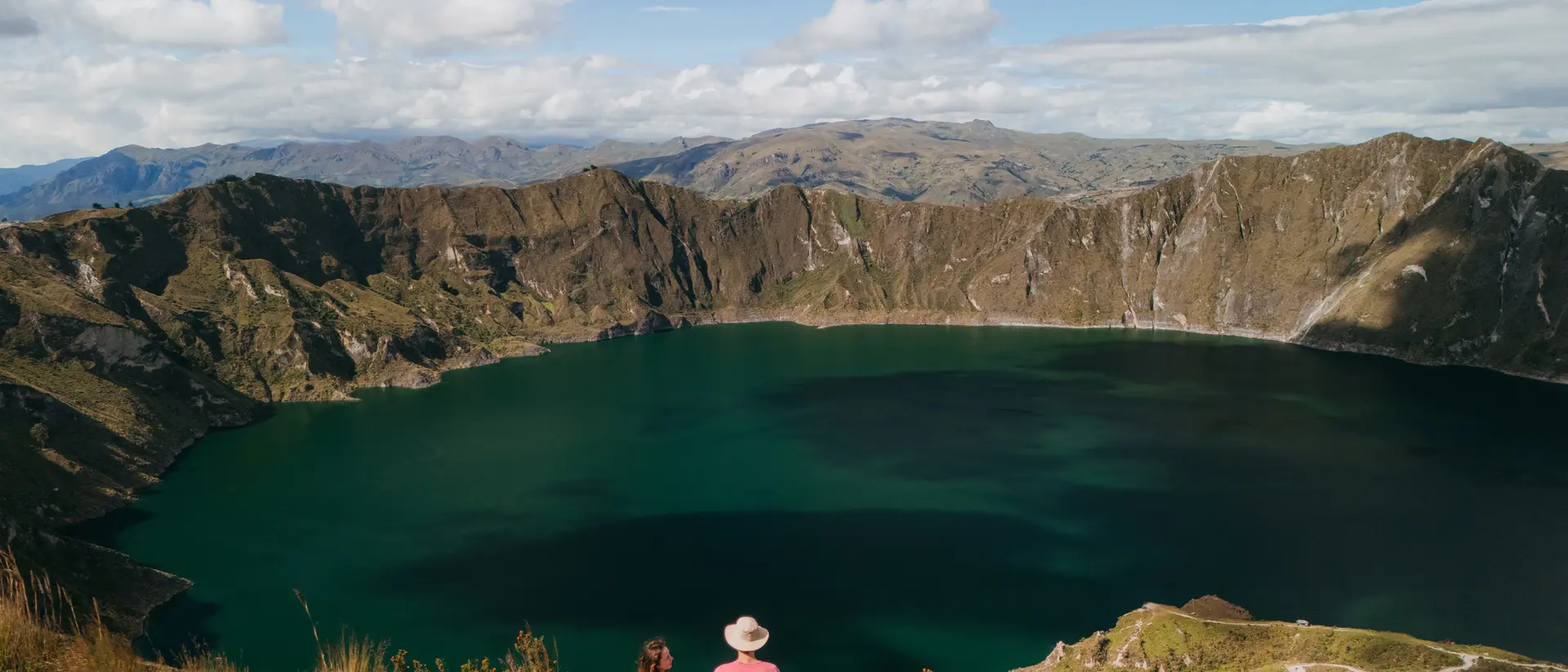 Two people stand on a hillside overlooking a large, serene lake surrounded by mountains under a blue sky with clouds.