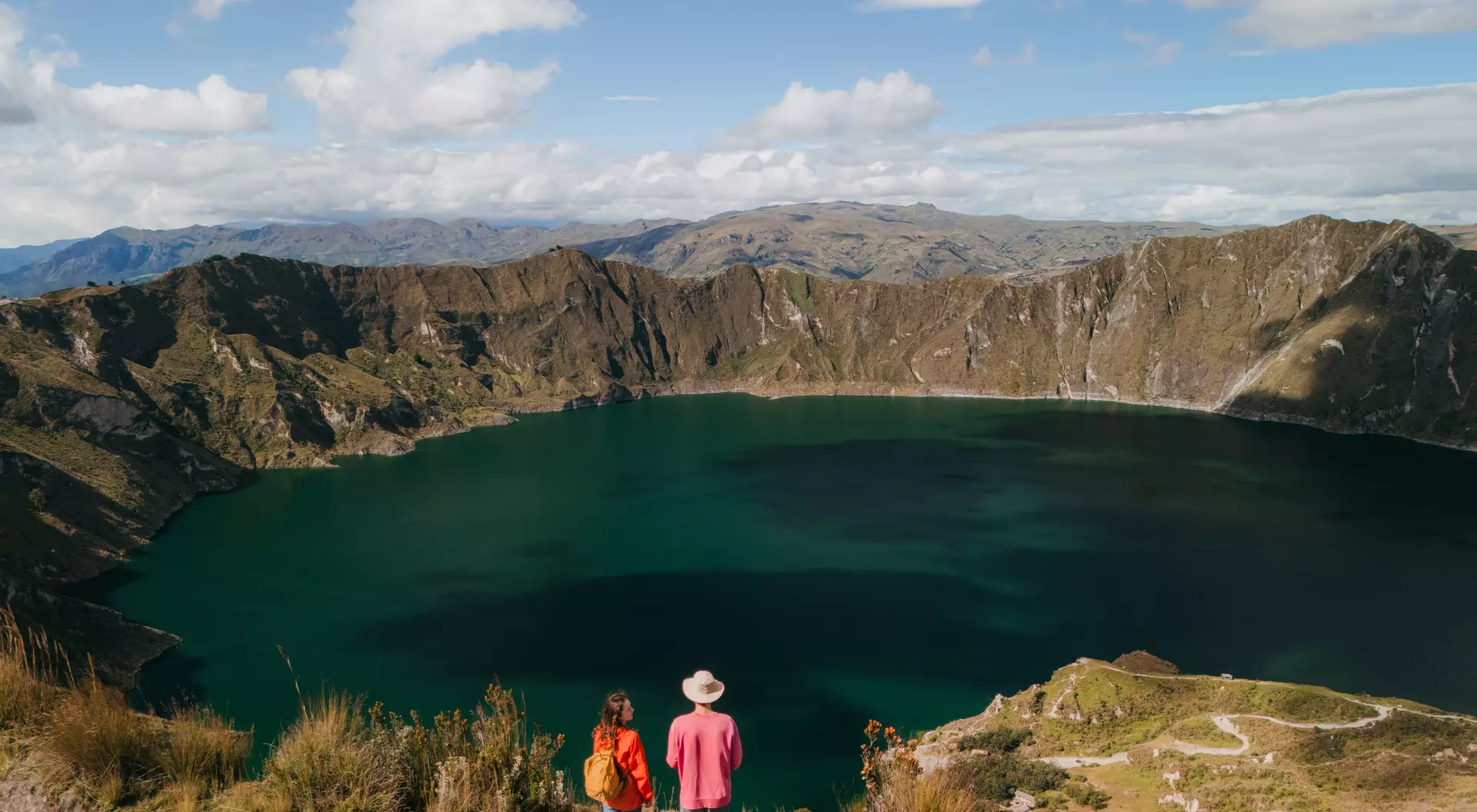 Two people stand on a hillside overlooking a large, serene lake surrounded by mountains under a blue sky with clouds.