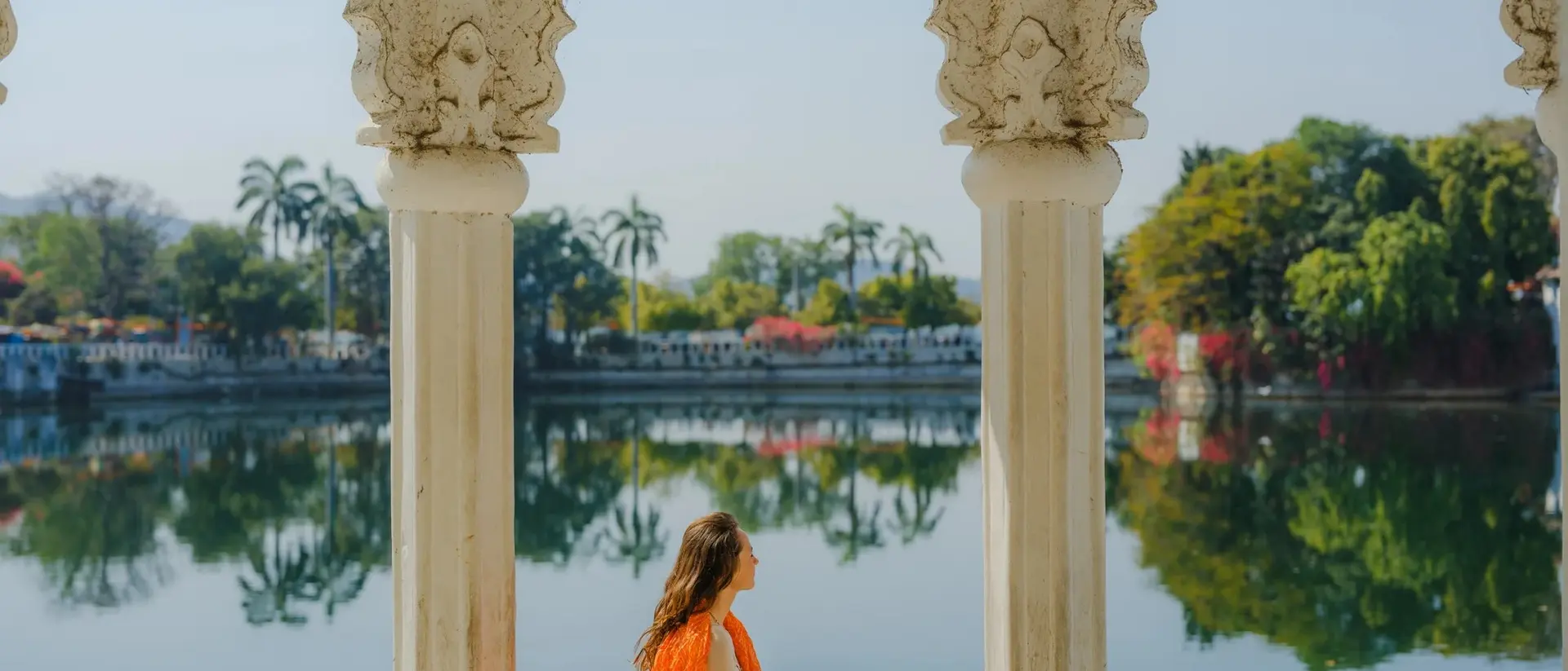 A woman in an orange outfit sits by a serene lake, framed by ornate white pillars and lush greenery in the background.