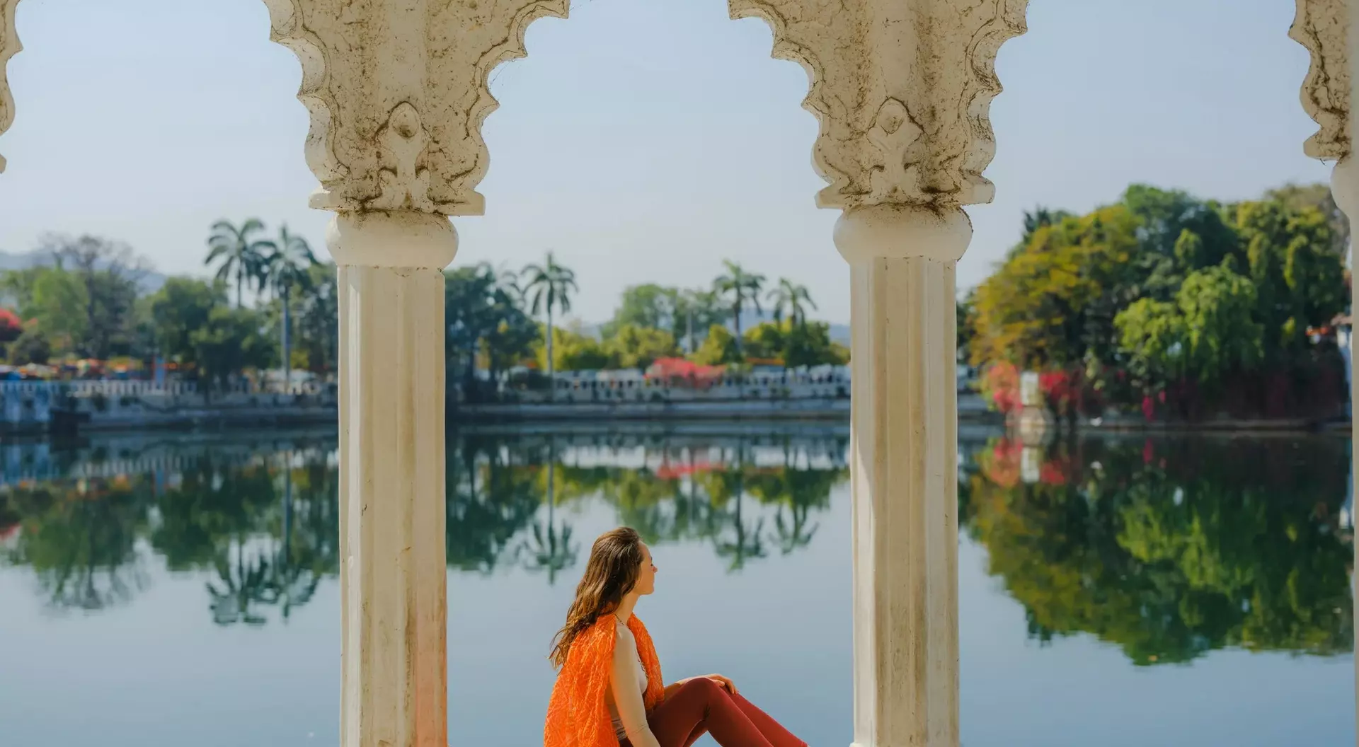 A woman in an orange outfit sits by a serene lake, framed by ornate white pillars and lush greenery in the background.
