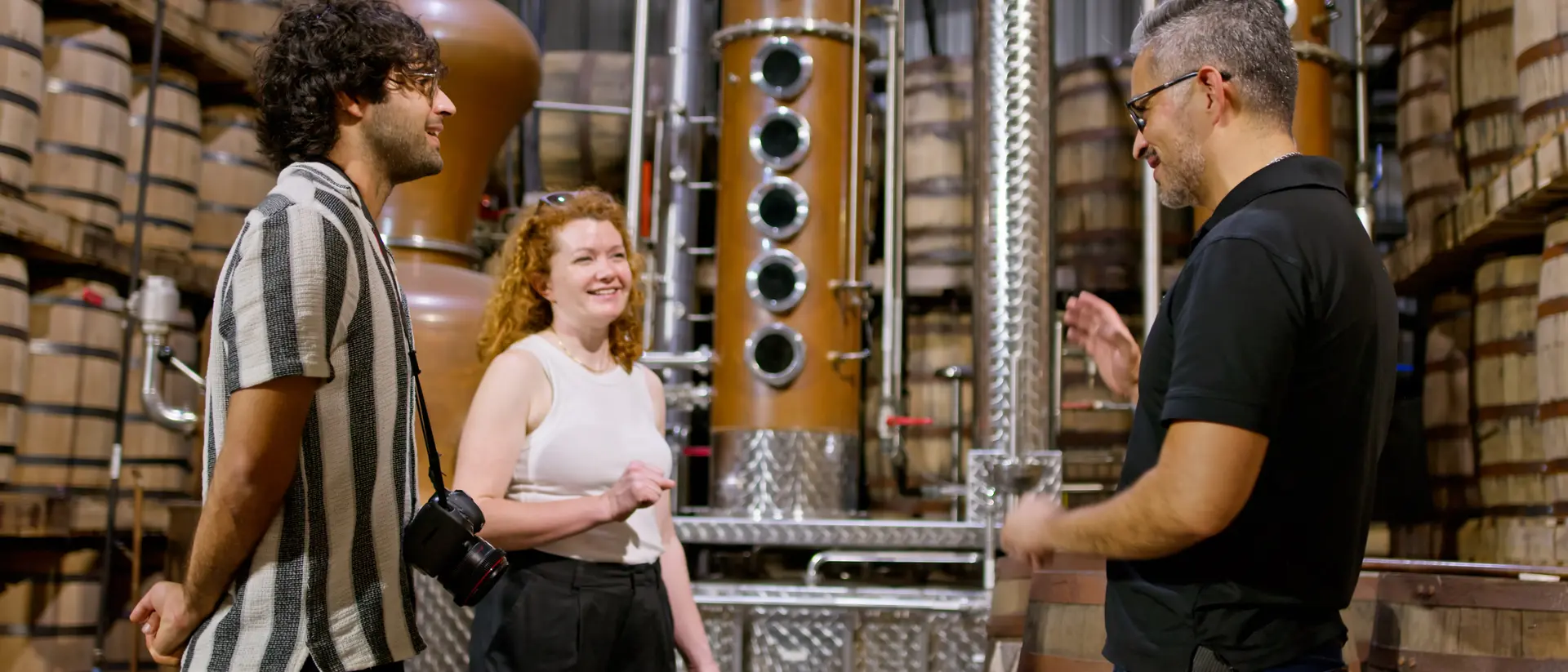 Two visitors smile and chat with a guide in a distillery surrounded by barrels and copper equipment.