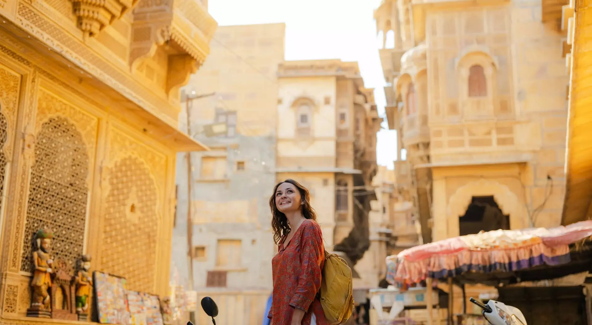 A woman in an orange dress stands in a sunlit narrow street, surrounded by historic buildings and parked motorcycles.