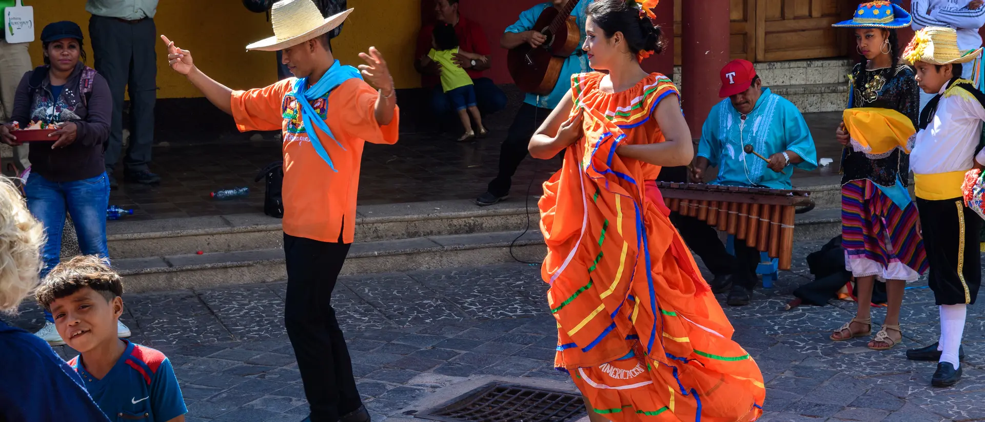 Colorful dancers perform a traditional dance in a lively outdoor setting, with musicians and a crowd around them.