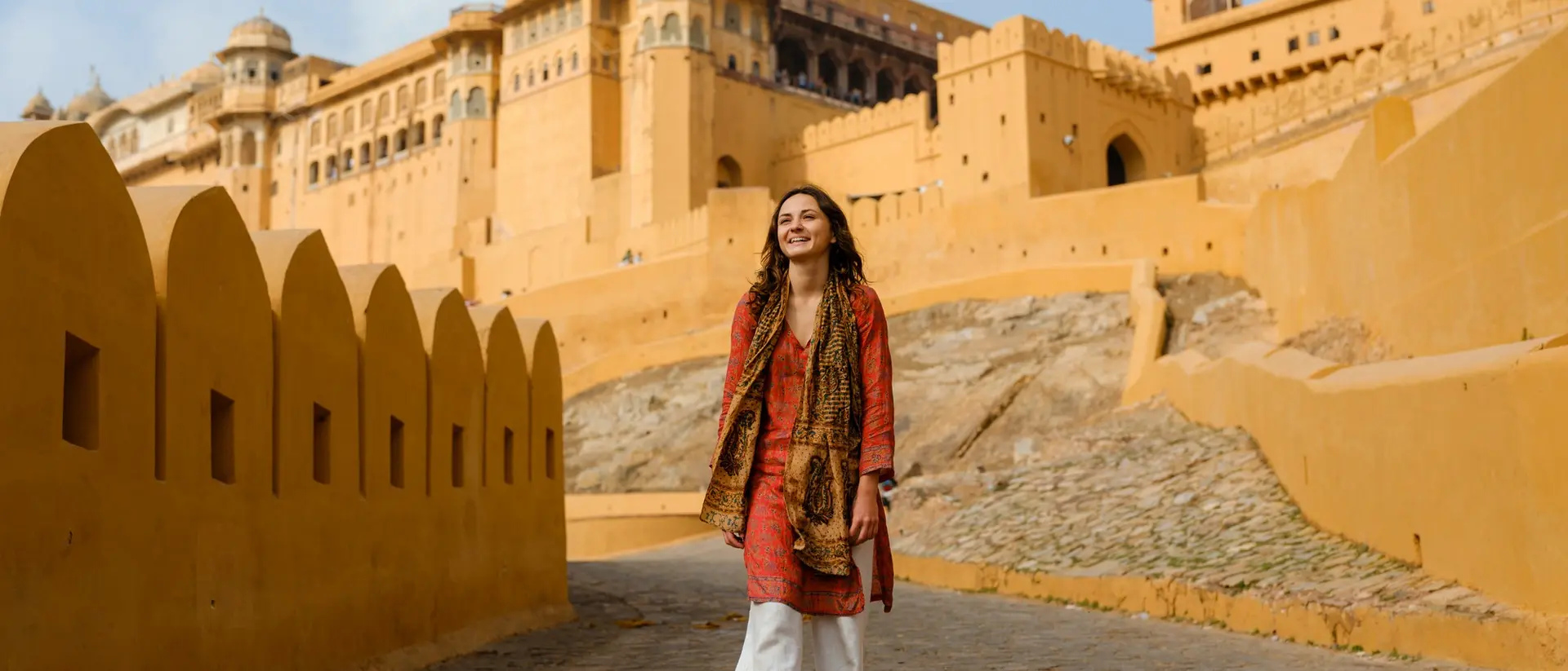 A woman walks along a stone path toward a large, orange fort against a blue sky.