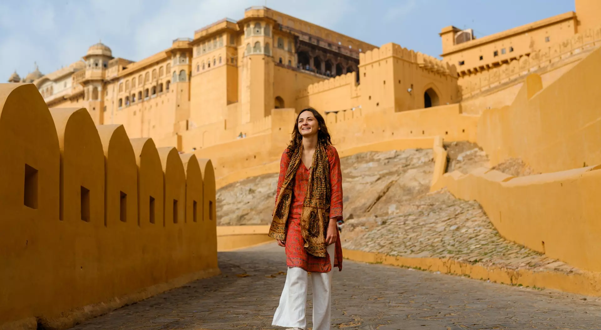 A woman walks along a stone path toward a large, orange fort against a blue sky.