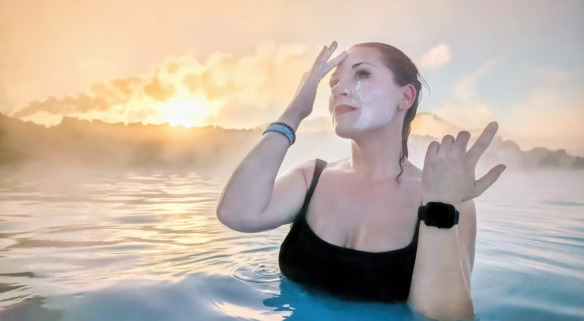 A person in a black swimsuit applies facial cream while standing in steam-filled water at sunset.