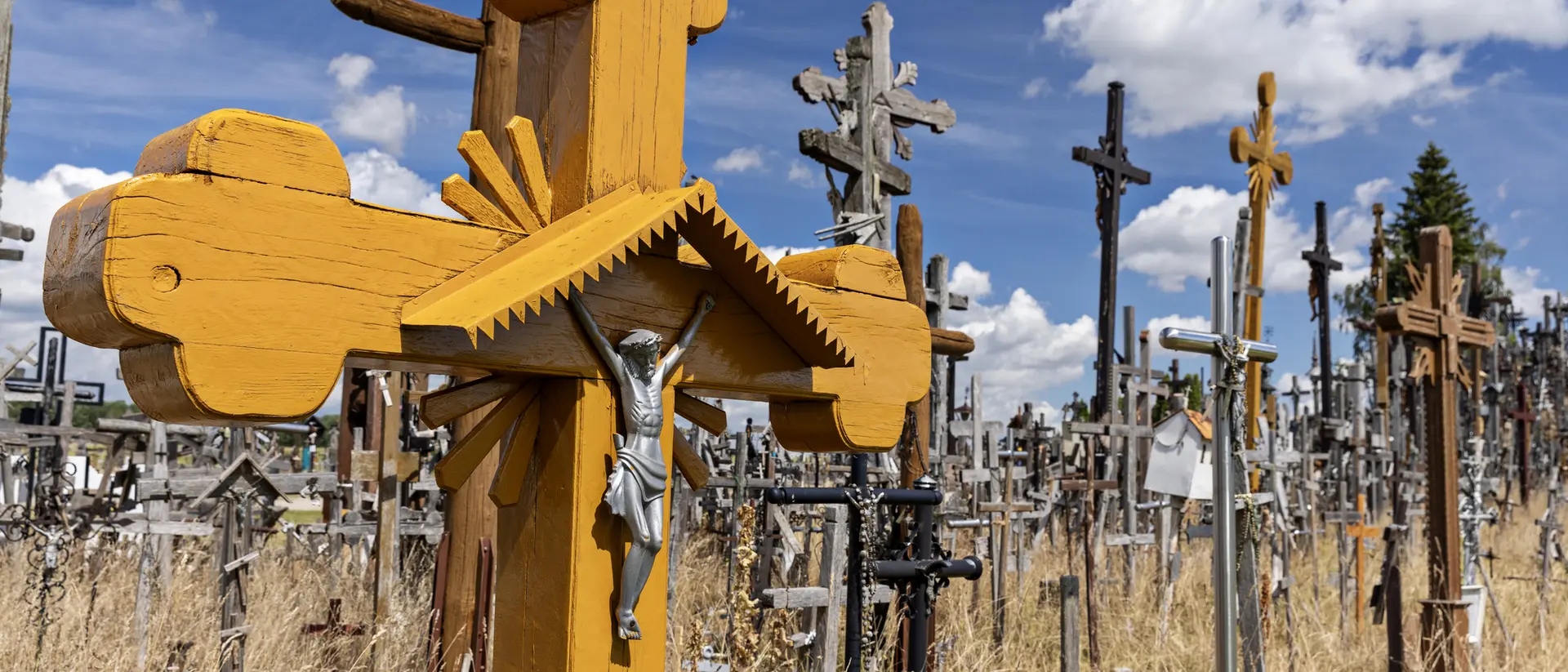 A wooden cross with a figure of Christ in the foreground, surrounded by numerous other crosses in a grassy cemetery.